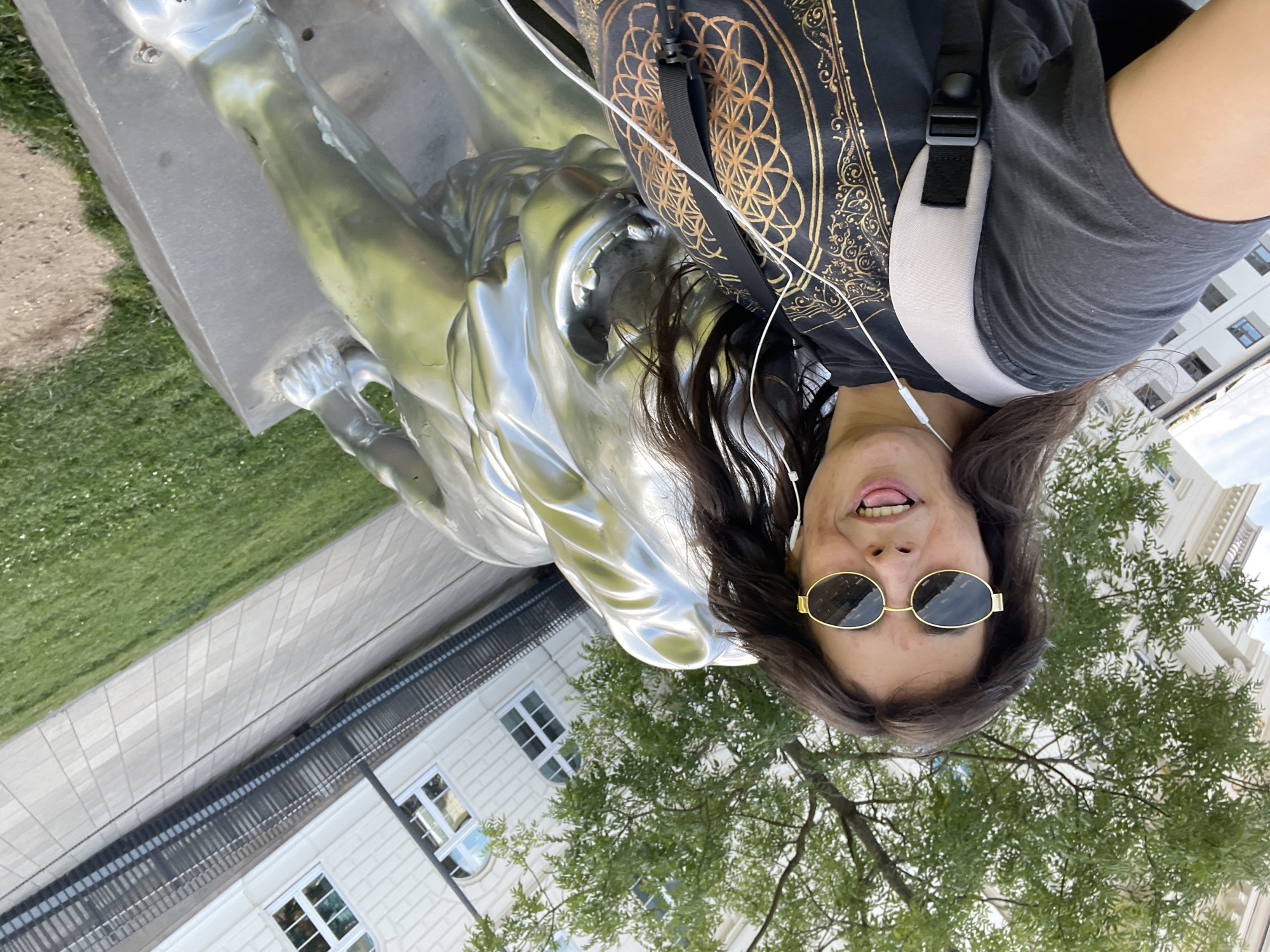 A young woman with long, dark hair is taking a selfie outdoors. She is wearing sunglasses, a dark t-shirt with a gold geometric design, and a light-colored backpack. She has earbuds in and her tongue is sticking out. Beside her is a large, silver sculpture of a lion. The sculpture is on a stone pedestal. The background shows a building with windows, a metal railing, trees, and grass. This image was taken in front of the main building of the University of Wuppertal.