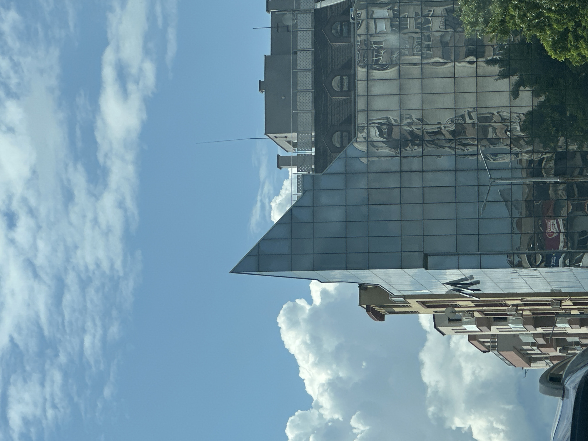 The image shows a view of buildings and the sky in Plovdiv, Bulgaria. A modern building with a triangular glass facade is prominent. The glass reflects other buildings, including one with arched windows, and nearby trees. The sky is blue with white clouds. At the bottom right, part of a car is visible, suggesting the photo was taken from inside a vehicle.