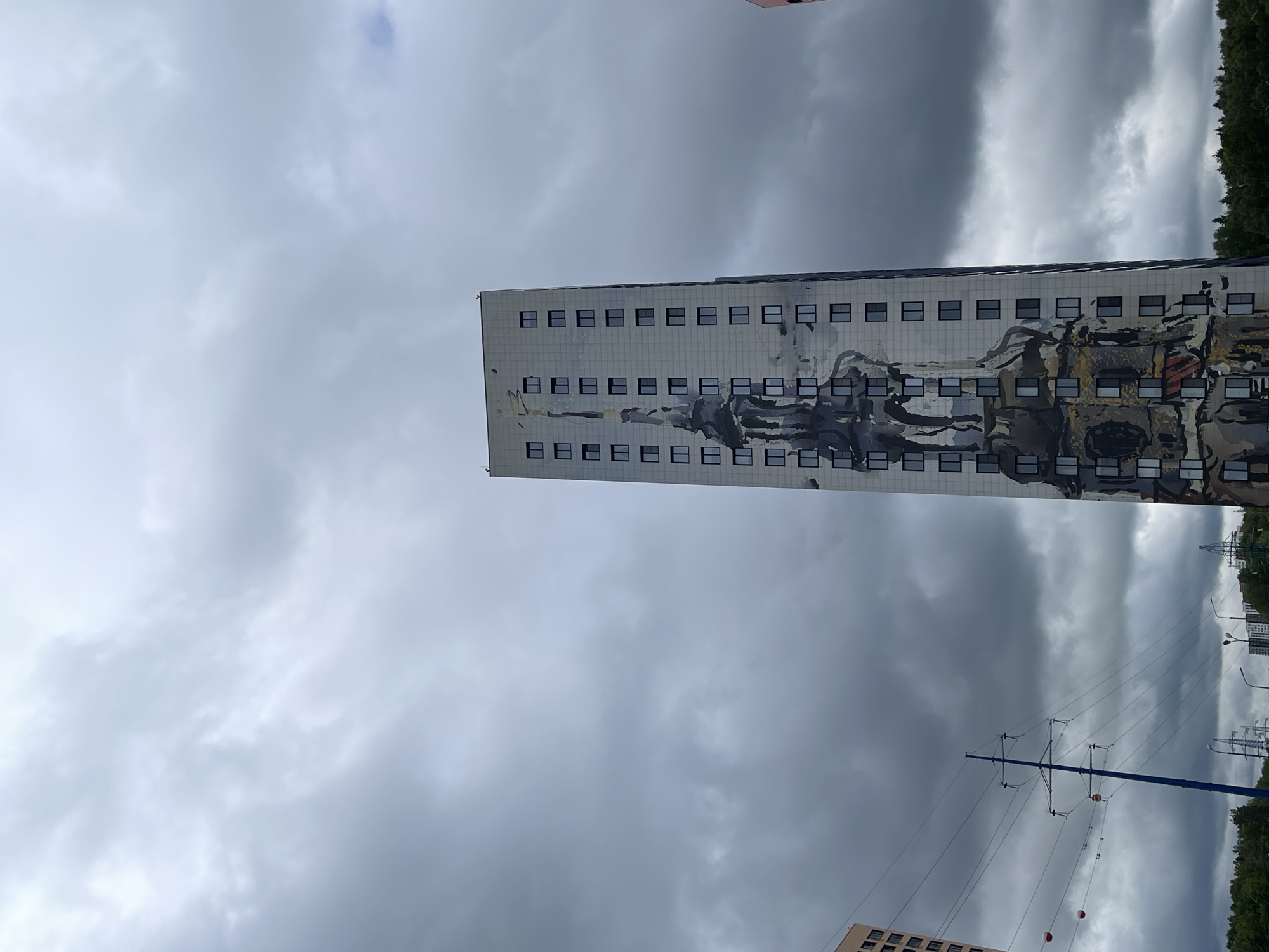 The image shows a tall building in Kommunarka, Russia, under a cloudy sky. The building has a rectangular shape with multiple rows of windows and a mural on its side. Power lines and poles are visible in the lower right of the image. Trees and other buildings are in the background.