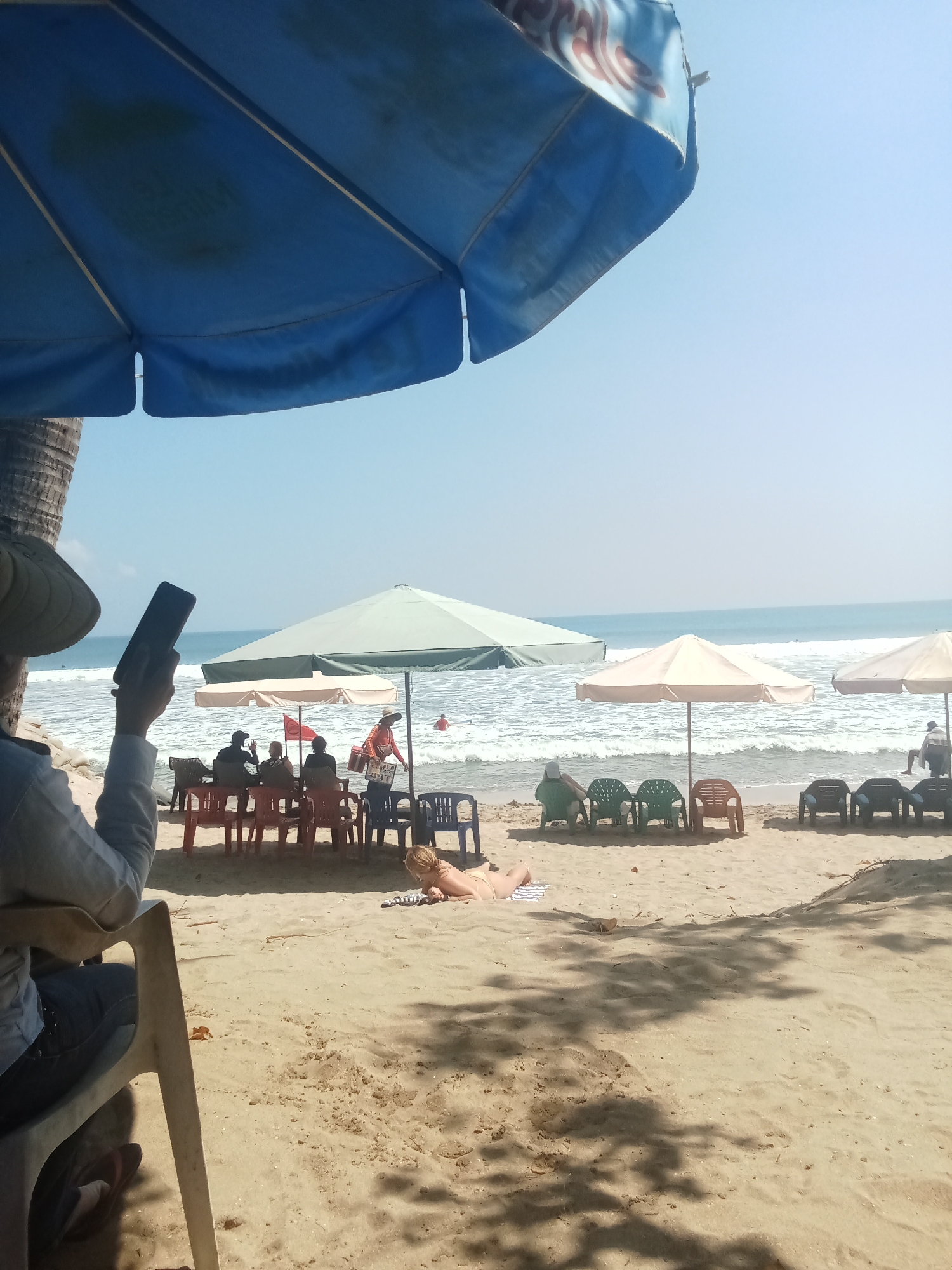 In the city of Unknown, Unknown, this image captures a sunny day at a beach. A large blue umbrella dominates the foreground, casting a shadow over the sandy expanse. Further back, several smaller umbrellas provide shade over rows of red and black plastic chairs, some occupied by silhouetted figures. A vendor in an orange shirt stands near the water's edge, holding a cooler. A woman in a yellow bikini lies on a towel in the sand, facing away from the camera. The ocean stretches to the horizon, with gentle waves lapping at the shore under a clear blue sky. In the lower left, a person wearing a wide-brimmed hat holds a dark rectangular object, likely a phone, in their right hand.
