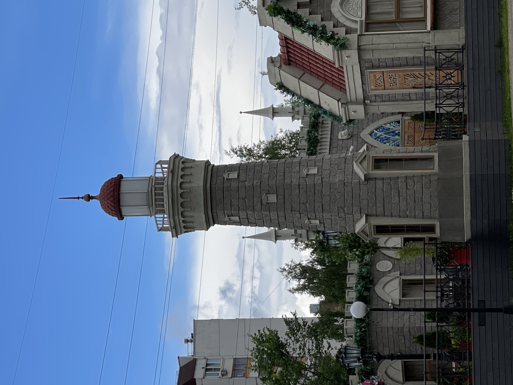 The image depicts a lighthouse-like tower and a building with castle-like architectural elements. The lighthouse has a red, conical roof with a spire on top and a white lantern room. The adjacent building is constructed of gray stone-like material, with decorative molding, arched windows, and red-tiled roofs. Greenery, possibly artificial vines, is draped along the building's facade. A black metal railing runs along a balcony or walkway. In the background, several tall, thin power lines crisscross the sky. A plain, modern building is visible behind the castle-like structure, and trees with lush green foliage are interspersed throughout the scene. The sky is blue with scattered white clouds, suggesting it is daytime. The overall impression is of a whimsical or themed architectural design, possibly for a commercial establishment or entertainment venue. No people or specific activities are evident.