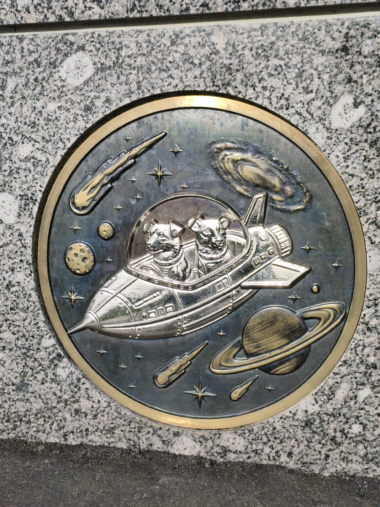 A circular, metallic relief plaque is embedded in a granite surface. The plaque depicts a space scene with two dogs in a rocket ship. The dogs are wearing astronaut helmets and appear to be piloting the ship. The background of the plaque features celestial bodies such as planets, stars, and a nebula, as well as comets. The rocket ship is detailed with windows and thrusters. The plaque has a bronze-colored border. The granite surface is speckled with gray, black, and white tones. The bottom of the image shows a dark, coarse surface, possibly pavement. This relief is located in Moscow, Russia, on the facade of a building at 18, Bolshaya Nikitskaya Street.