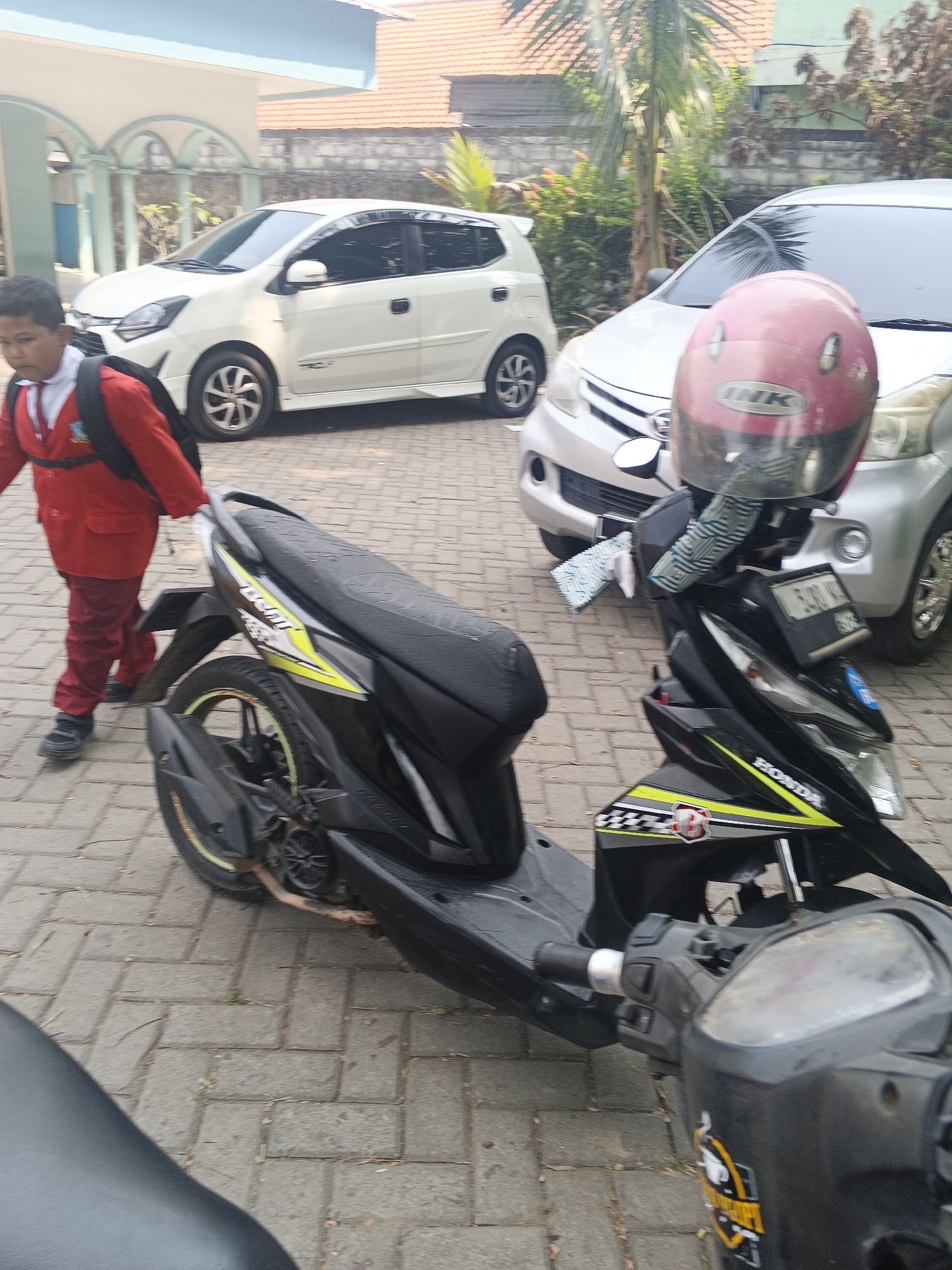In Krian, Indonesia, a young student in a red uniform with a black backpack is walking past a parked black scooter. The scooter has a pink helmet on its handlebars. Behind the scooter, a silver car is visible, and to the left, a white car is parked. The scene appears to be on a paved area, possibly a school or public space, with buildings and greenery in the background.