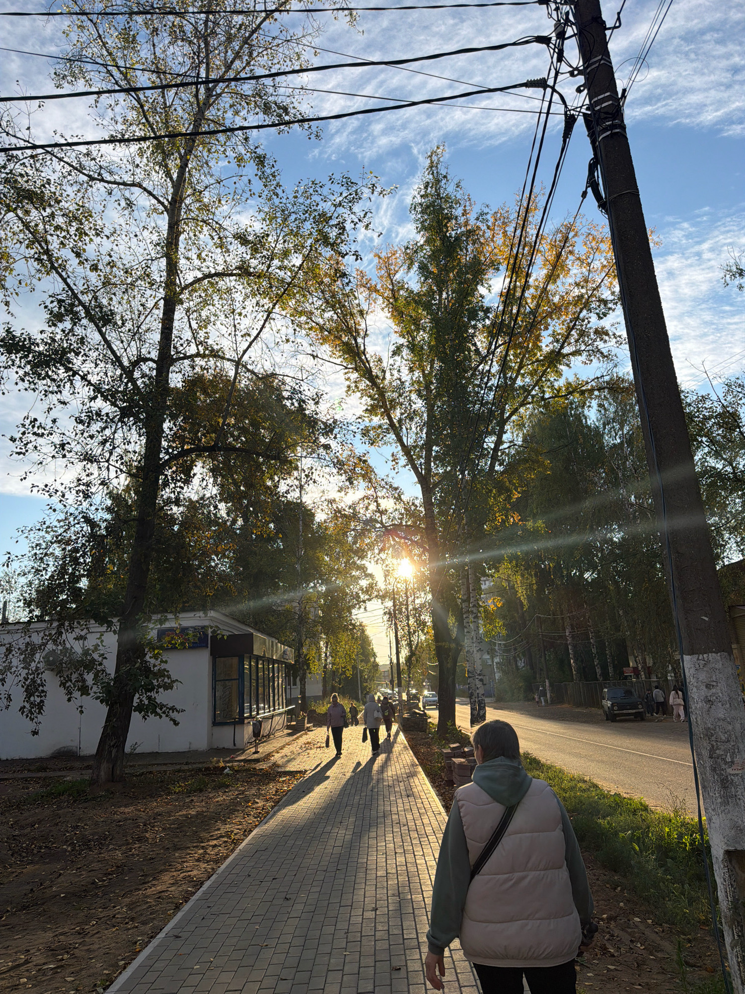 The image depicts a sidewalk scene in Chaikovsky, Russia, with a paved walkway in the foreground leading away from the viewer. A person, wearing a light-colored puffer vest over a green hooded sweatshirt and dark pants, walks away from the camera. To the left of the sidewalk, there is a white building with a dark-framed window and an awning. Bare trees with some remaining autumn leaves cast long shadows across the walkway. Power lines stretch across the sky, supported by a utility pole on the right side of the frame. Beyond the building, a road is visible with a few cars and more trees lining the other side. The sun is low in the sky, creating a warm, golden light and lens flare. Several other individuals are walking on the sidewalk further ahead, walking towards the viewer. The overall environment appears to be an urban or suburban street during autumn.