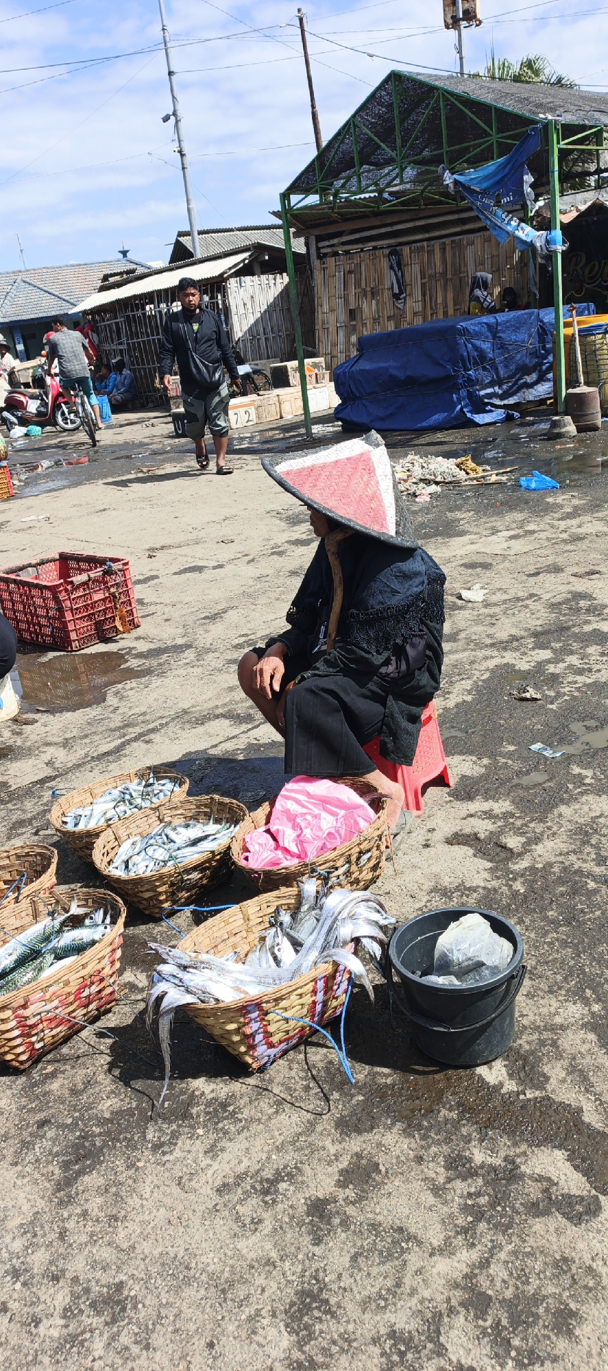 A vendor wearing a conical hat squats beside several woven baskets filled with fish at an outdoor market, likely in Puger, Indonesia. The vendor is dressed in black. In front of the vendor are three baskets containing small fish, and one basket containing a pink plastic bag. To the right of the baskets is a black bucket containing a large grey object. In the background, a man in a black jacket and grey pants walks away from the camera, and other people and structures are visible. The ground is made of concrete and appears wet in places. The sky is blue with some clouds.