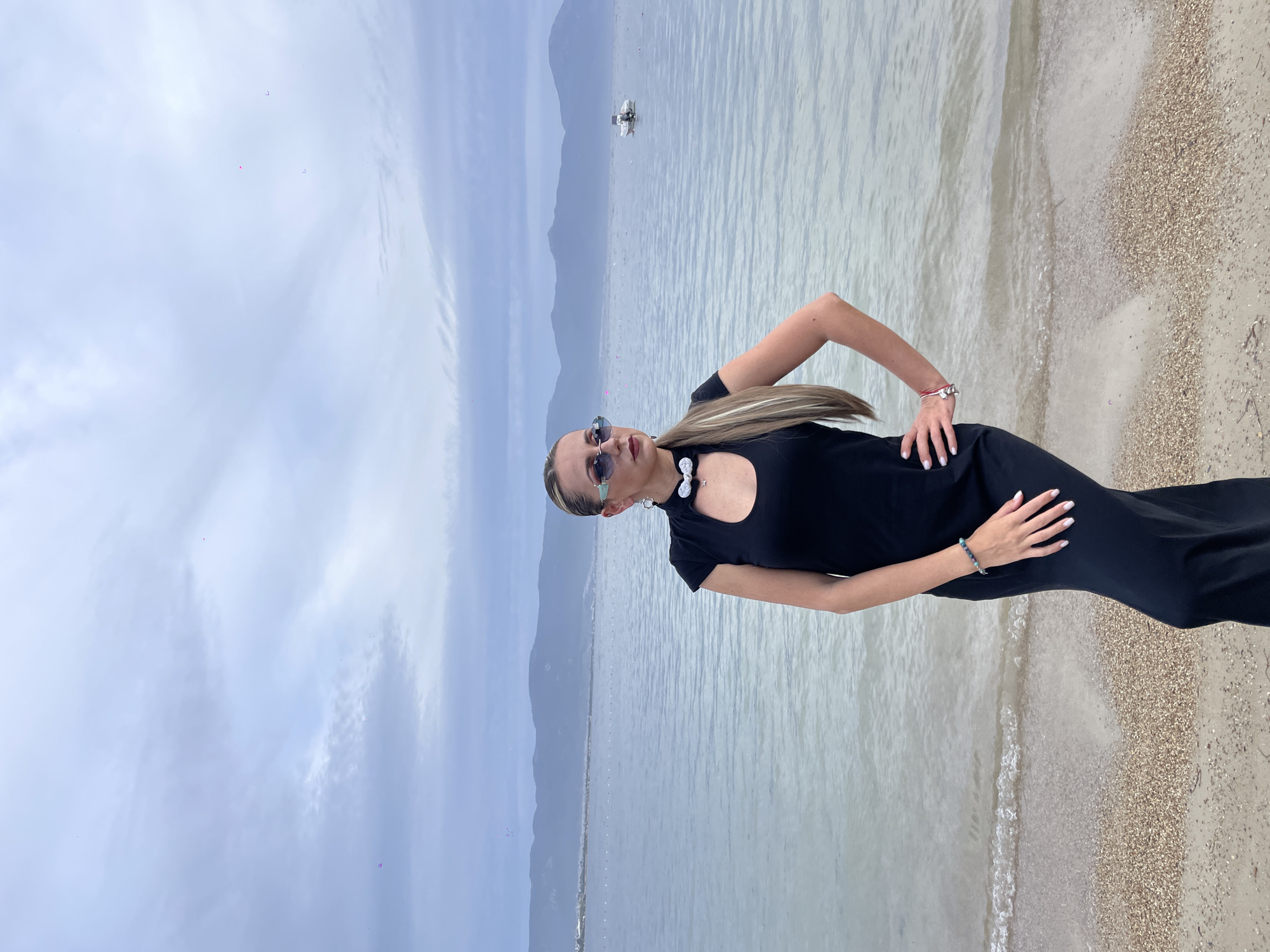 A vertical medium shot depicts a woman standing on a beach in Kuşadası, Turkey. She is wearing a black dress, sunglasses, and jewelry. Her hands are on her hips, and she appears to be posing for the picture. The sea is behind her, and mountains are visible in the distance. There is a boat on the water. The sky is cloudy. The beach is sandy with small stones.