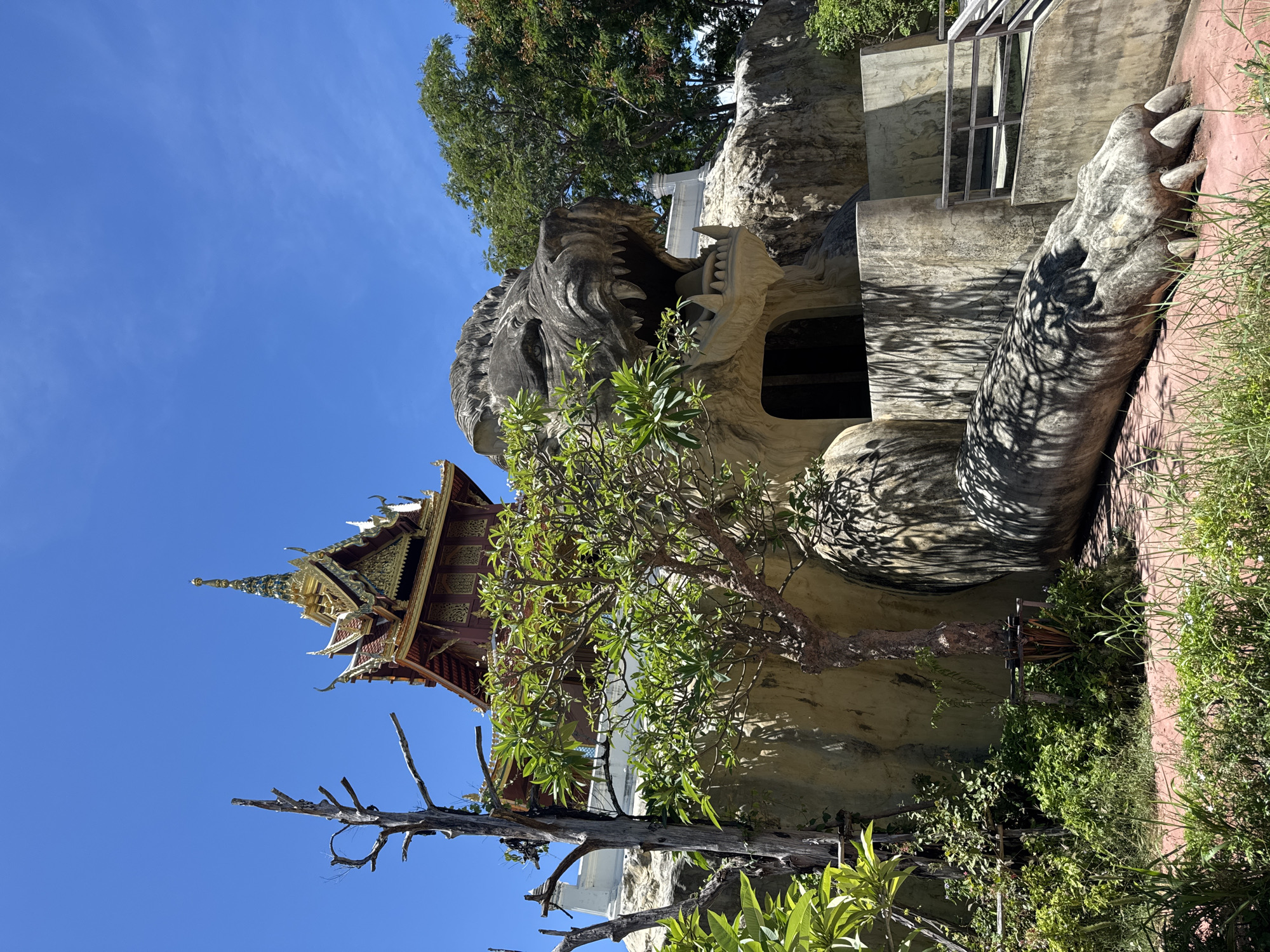 The image captures a unique architectural structure in Bang Pu Subdistrict Municipality, Thailand. A large, imposing sculpture of a lion's head serves as the entrance to a building, with its open mouth leading into a dark interior. A massive leg and claw extend from the side of the structure. Adjacent to the lion sculpture, a traditional Thai building with a multi-tiered roof and intricate golden details is visible. A tree with green leaves partially obscures the view, while a dead, leafless tree stands closer to the viewer. The sky is a clear blue.