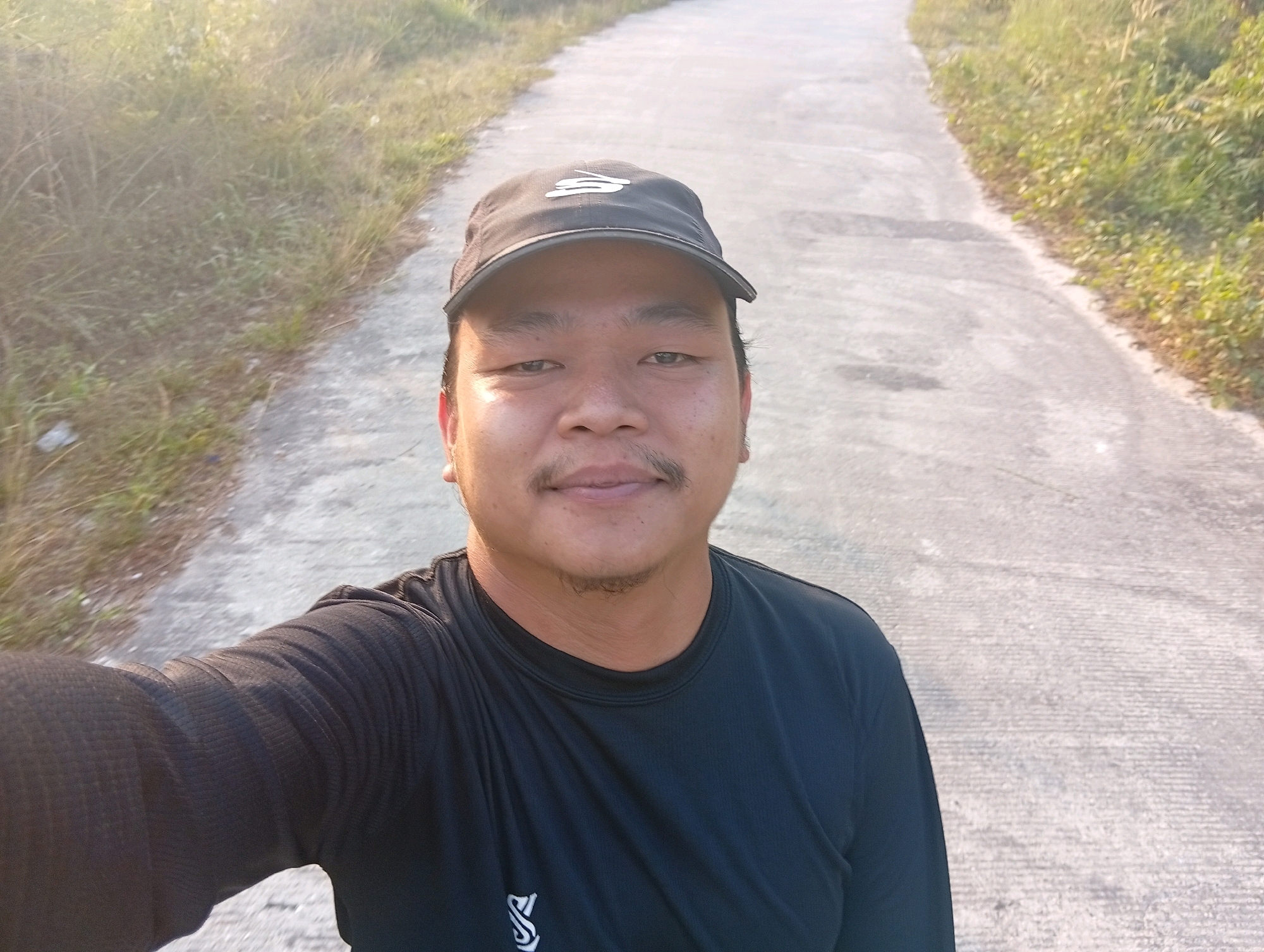 In Kualu, Indonesia, an individual is captured in a selfie as they stand on a concrete path bordered by dry grass and vegetation. The person, wearing a black baseball cap and a black long-sleeved shirt, is looking directly at the camera with a slight smile. The lighting suggests it is either early morning or late afternoon, casting a warm glow on the scene. The background shows a seemingly rural or suburban road stretching into the distance, with greenery on either side. The overall impression is of a casual outdoor moment.
