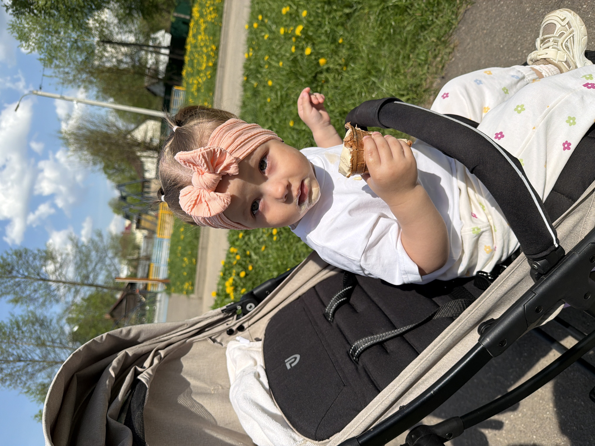 A young child sits in a stroller, holding and eating from a cone, with food smeared around their mouth. The child wears a white t-shirt, patterned pants, and a pink headband. The stroller is beige and black. The background shows a grassy area with yellow flowers, trees, and a blue sky. The location is potentially Reshetnikovo, Russia.