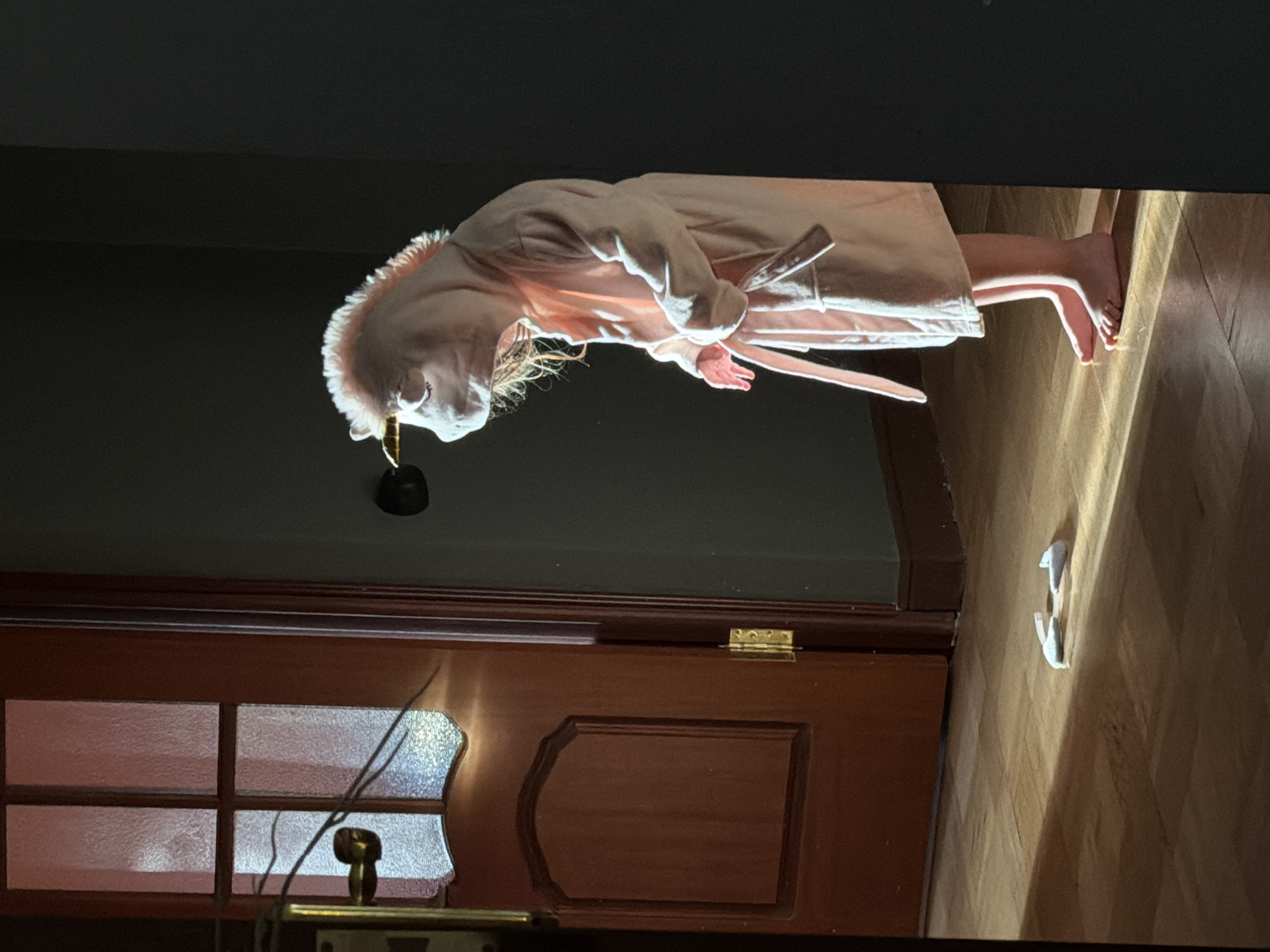A person wearing a unicorn costume and a pink robe stands on a wooden floor in a dimly lit room. The unicorn costume covers their head and torso, with a golden horn protruding from the forehead and a pink mane. The robe is tied at the waist with a belt. The person is looking down, with their hands clasped in front of them. Two white shoe inserts lie on the floor in the foreground. To the left, a wooden door with frosted glass panels is visible, with a brass doorknob and hinges. The scene is illuminated by a shaft of sunlight that casts long shadows across the floor. The specific location within the city of Khimki, Russia cannot be determined from the image.