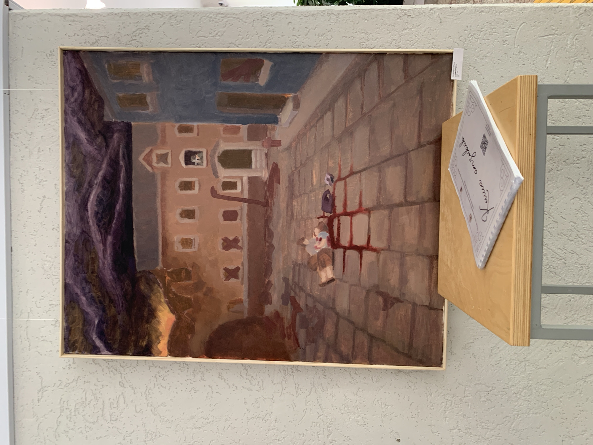 A horizontally-oriented photograph depicts a painting exhibited indoors. The painting, oriented vertically, features a brown building on the left side with multiple windows and a gray roof. The building is adjacent to what appears to be a dark purple or gray tree trunk. On the right side of the painting is a tan-colored cobblestone street. Two figures are visible on the street. Red streaks are painted on the cobblestone, suggesting the aftermath of violence or an injury.
The painting hangs against a textured, off-white wall. A small, light-colored wooden platform is attached to the right side of the painting's frame. A white paper containing writing is placed on the wooden platform. A silver-colored metal frame is located to the right of the wooden platform.
The location is Chaikovsky, Russia.