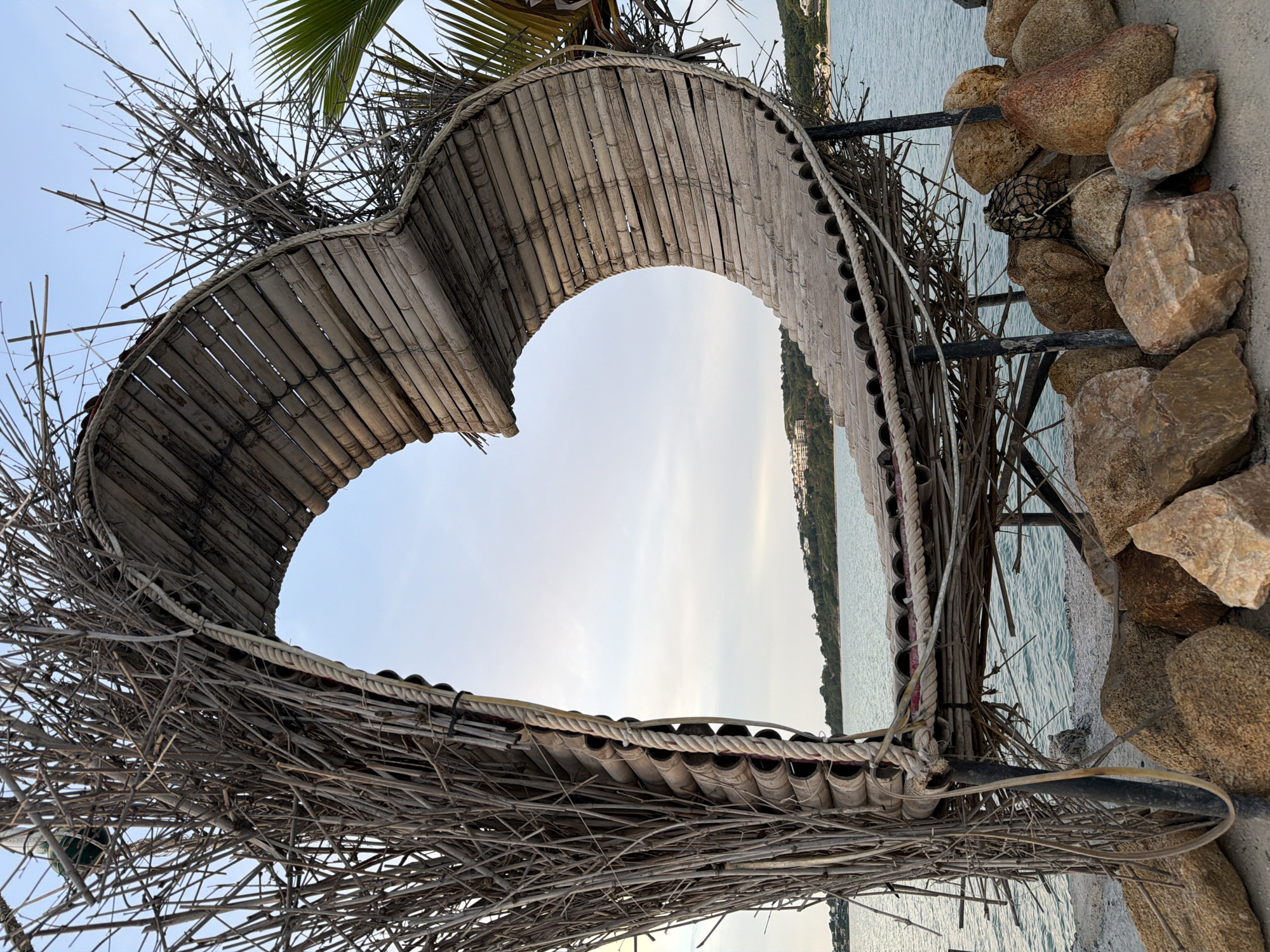 An outdoor, eye-level, full shot captures a large, heart-shaped sculpture made of bamboo and branches situated on a rocky shore with a body of water and distant landscape behind it. The sculpture, likely intended as a photo opportunity, is constructed from split bamboo segments that form the curving outer edge of the heart, bound together with rope. The interior of the heart is filled with a dense arrangement of dried branches and twigs, creating a natural, rustic aesthetic. The sculpture is supported by dark metal poles, partially obscured by the rocks at its base.
The foreground is dominated by a collection of large, rough-hewn rocks of varying sizes and colors, ranging from light tan to reddish-brown, resting on a sandy or gravelly surface. The body of water in the mid-ground is a calm, turquoise blue, with small ripples visible on its surface. Beyond the water, a lush green hillside is dotted with buildings, suggesting a populated coastal area. The sky above is a pale blue with wispy white clouds, indicating either early morning or late afternoon light. A frond of a palm tree is visible in the upper right corner, adding to the tropical feel. The overall scene conveys a serene and picturesque coastal setting in the Ko Samui District of Thailand.