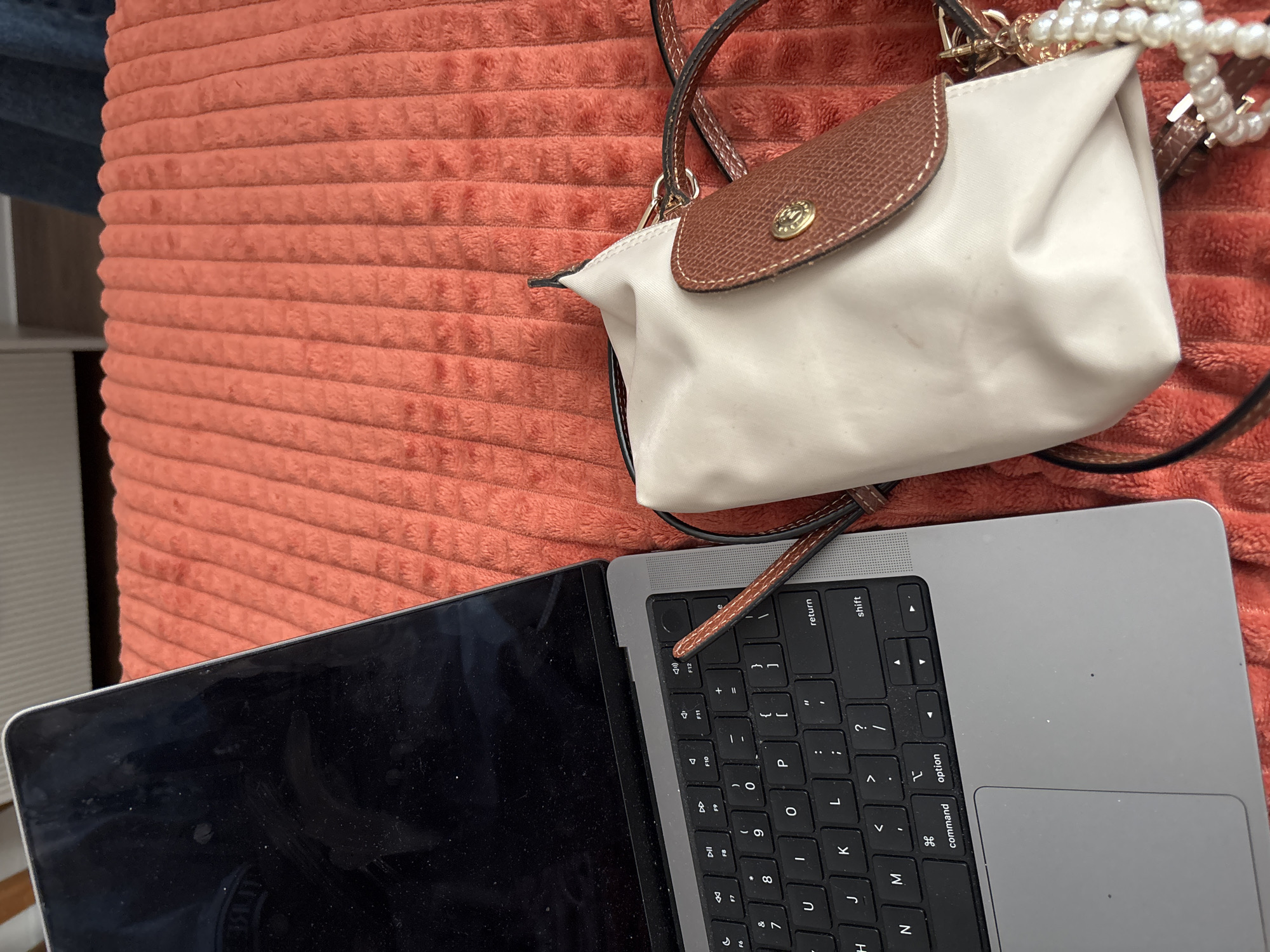A close-up shot displays a laptop, a handbag, and a textured surface. The laptop, positioned at the bottom left, features a dark screen and a gray keyboard. Adjacent to it is a white handbag with brown leather accents and pearl embellishments. The backdrop is a coral-colored, horizontally ribbed, textured surface.