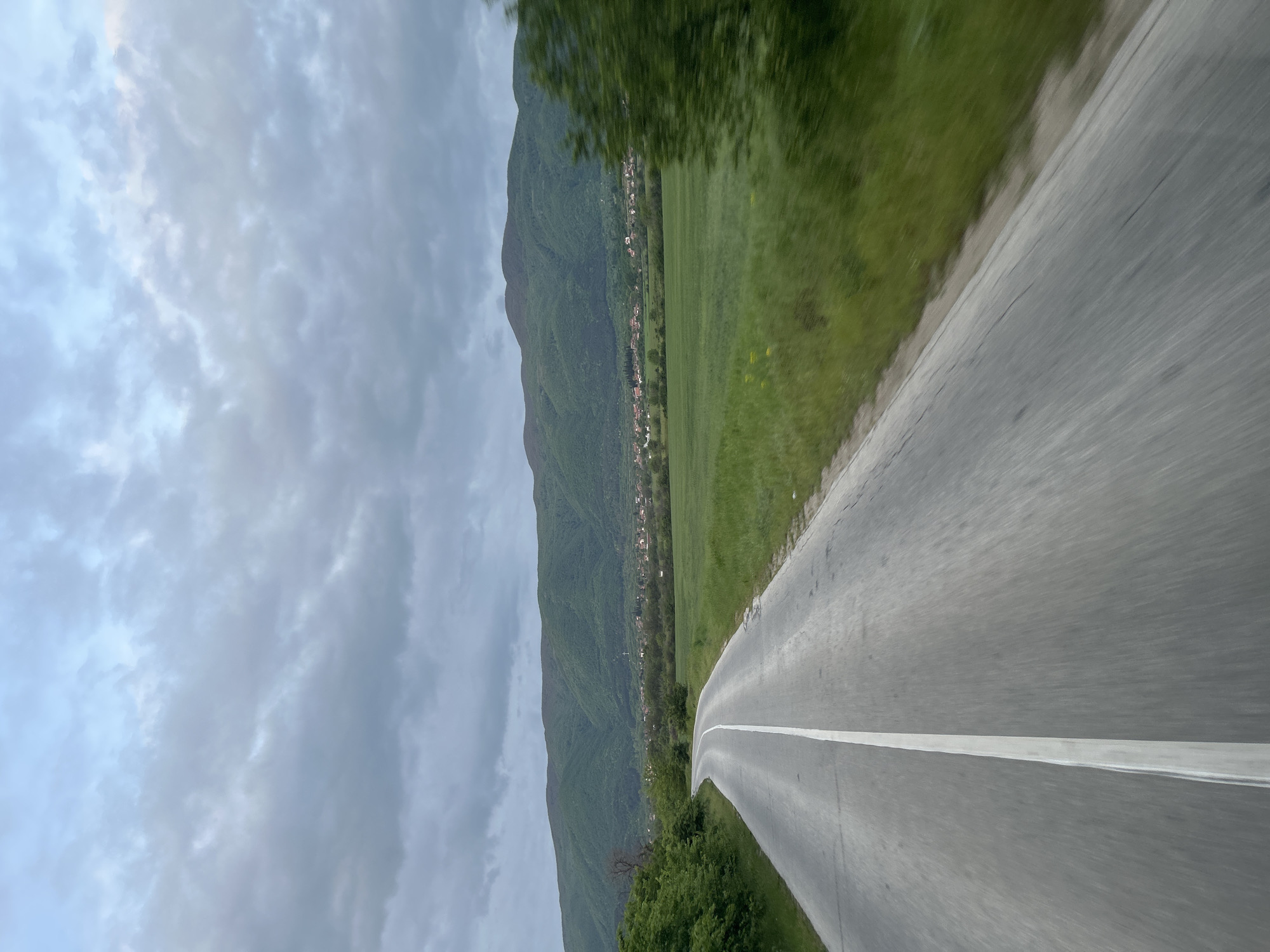 The image shows a view from a road in Patalenitsa, Bulgaria. The road is gray asphalt with a white center line and stretches into the distance. Adjacent to the road is a green field. In the background is a forested mountain range with a village nestled at its base. The sky is cloudy.