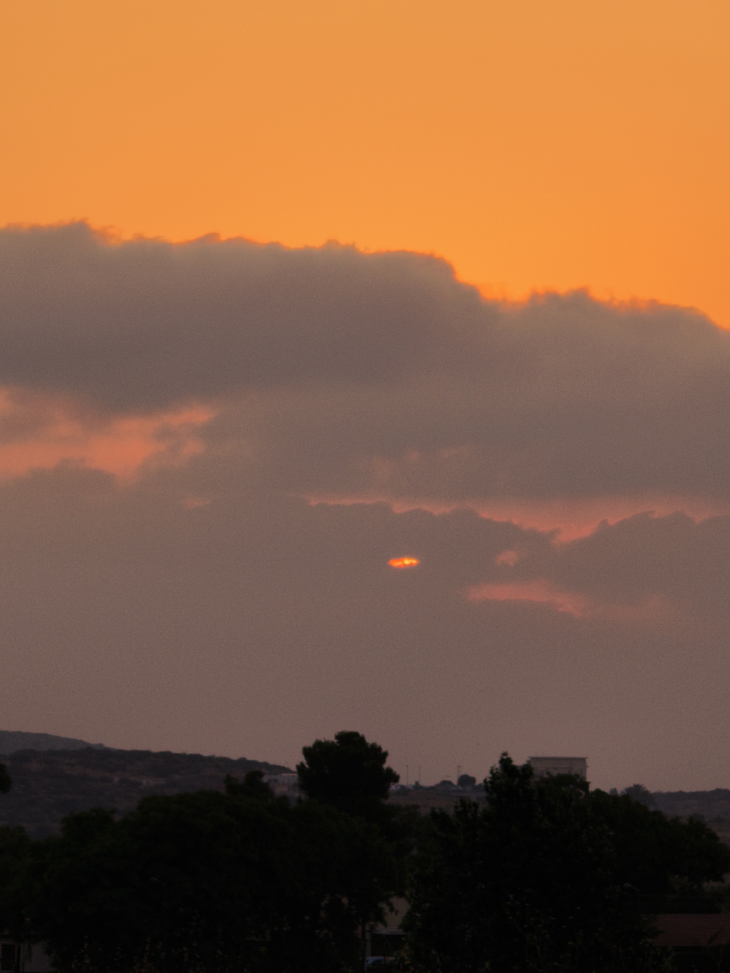 A sky filled with orange hues at sunset provides the backdrop for a hazy landscape. Dark clouds are present in the middle ground, with a small, bright orange light visible through a gap in the clouds, possibly a glimpse of the setting sun. In the foreground, the silhouettes of trees and a distant hill with some structures are visible. The scene suggests a location in Lachish Regional Council, Israel, during twilight.