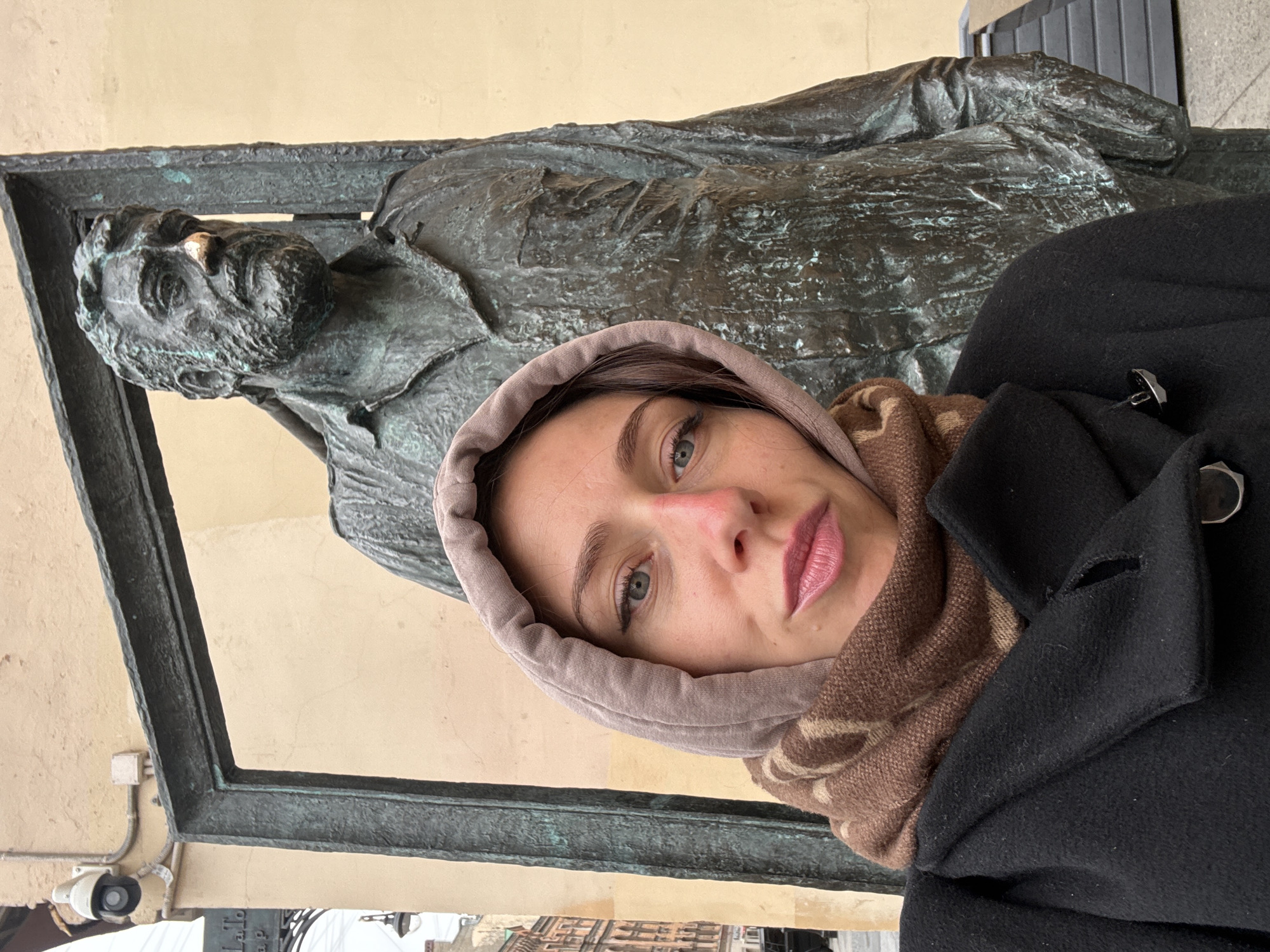 A woman in a light brown hoodie, patterned brown scarf, and a black coat is positioned to the right and in front of a bronze relief of Fyodor Dostoevsky. The relief depicts a reclining Dostoevsky within a dark bronze frame. The relief is mounted on a pale yellow wall. A surveillance camera is visible near the bottom left. The location is likely Saint Petersburg, Russia, due to Dostoevsky's association with the city and the architectural style of the buildings visible in the background. The woman is facing towards the camera.