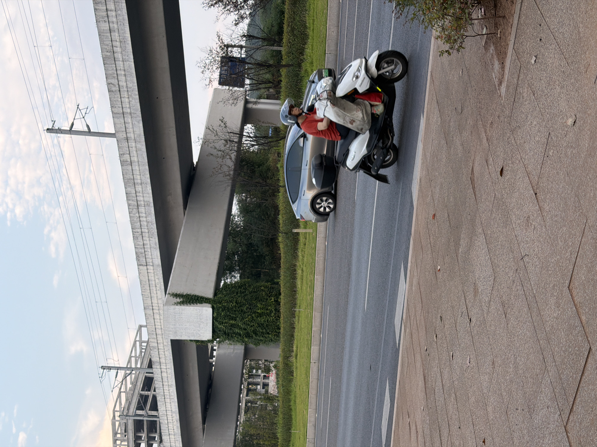 A person wearing a red shirt and a helmet is riding a white scooter on a road in Jiangjin, China. The scooter has a white bag draped over the handlebars. Behind the scooter, a silver car is visible. In the background, a large concrete overpass with overhead power lines runs across the scene. The overpass is supported by a pillar covered in green ivy. A section of a building under construction is also visible to the left. The road is paved and has lane markings. A sidewalk made of stone tiles is in the foreground. Trees and vegetation line the roadside. The sky is overcast with a few clouds.