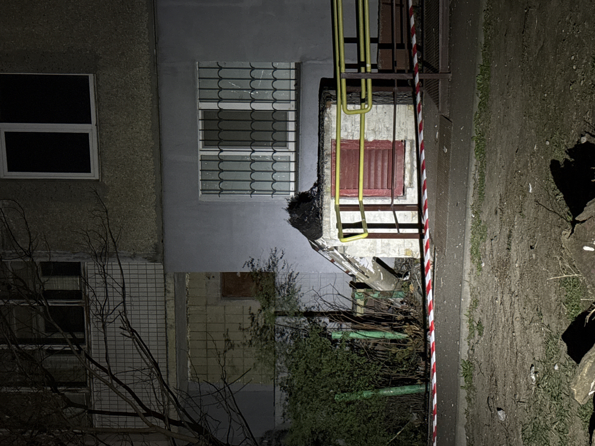 The image shows the aftermath of an explosion at a building in Kyiv, Ukraine. The building has visible damage, including broken windows and debris scattered around. A section of the building has collapsed, and the debris is marked with red and white tape. The scene appears to be at night, illuminated by artificial light. There are bare trees and shrubs in the foreground.