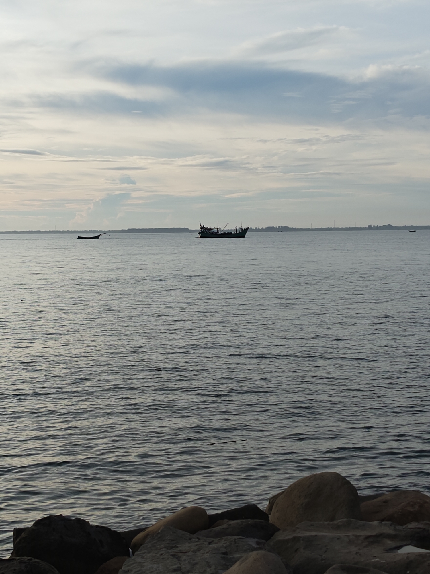 The image depicts a seascape with several boats on the water, taken in Lhokseumawe, Indonesia. In the background, a coastline is visible, suggesting land across the water. The sky is overcast with varying shades of grey and white clouds. The water is relatively calm, with small ripples across the surface. In the foreground, there are rocks that make up a shoreline. Two boats are visible in the distance, one larger and more detailed than the other.