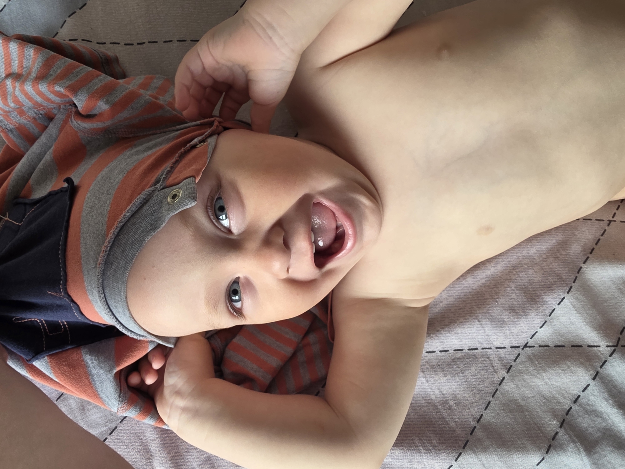 A baby or toddler is captured in a close-up, high-angle shot, lying on their back indoors. The child's head is tilted, and they are looking directly at the camera with wide, expressive blue eyes. Their mouth is open wide, revealing their tongue and gums, suggesting they are mid-laugh, babble, or making a sound, conveying a joyful and playful emotion.
The baby has fair skin and is partially clothed in a long-sleeved garment with alternating orange-red and gray horizontal stripes. Parts of their chest and belly are bare. One of their hands is raised near their head, seemingly touching or adjusting the striped fabric, while the other hand is lower, resting on their side.
The background is a light-colored, possibly gray or beige, soft fabric, likely a bed sheet or blanket, adorned with a pattern of dashed black lines forming diamonds or a grid. The lighting is bright and even, consistent with an indoor setting during the daytime. No specific location cues are visible within the frame, but the context indicates the image was taken in Chaikovsky, Russia. There is no visible text in the image.