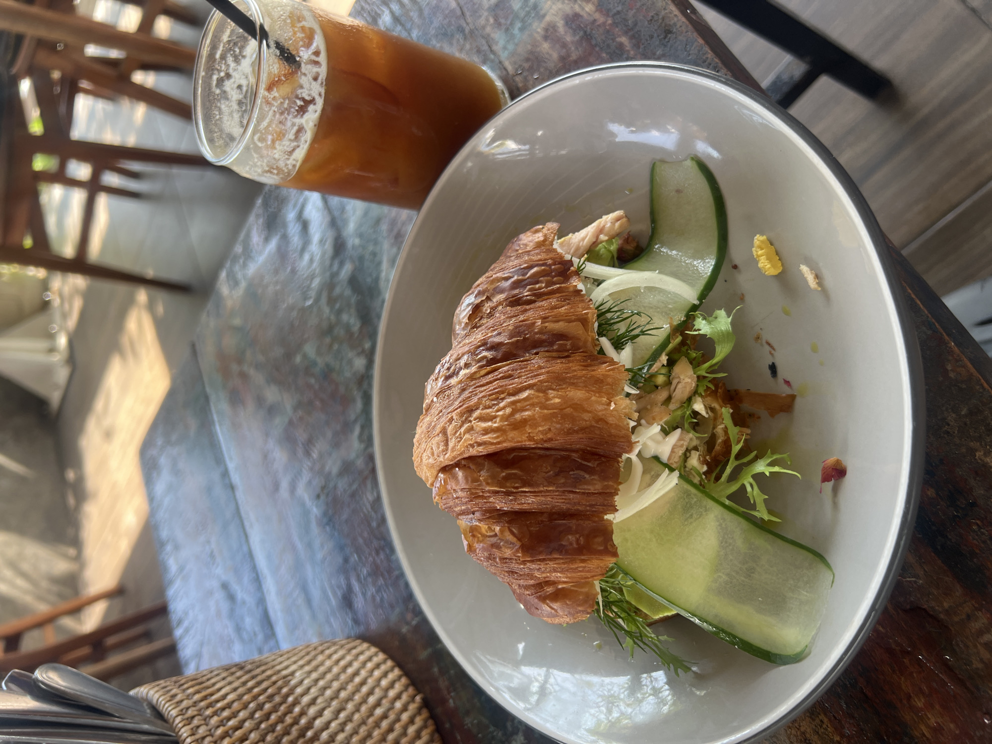 The scene is a cafe in North Kuta, Indonesia. A plate containing a croissant with salad is on the wood table, accompanied by a glass of iced tea. The croissant is light brown with layers of pastry visible. The salad includes sliced cucumbers, lettuce, and white shredded vegetables. The iced tea is light brown and has a straw. The table shows some wear and tear. A chair can be seen in the background.