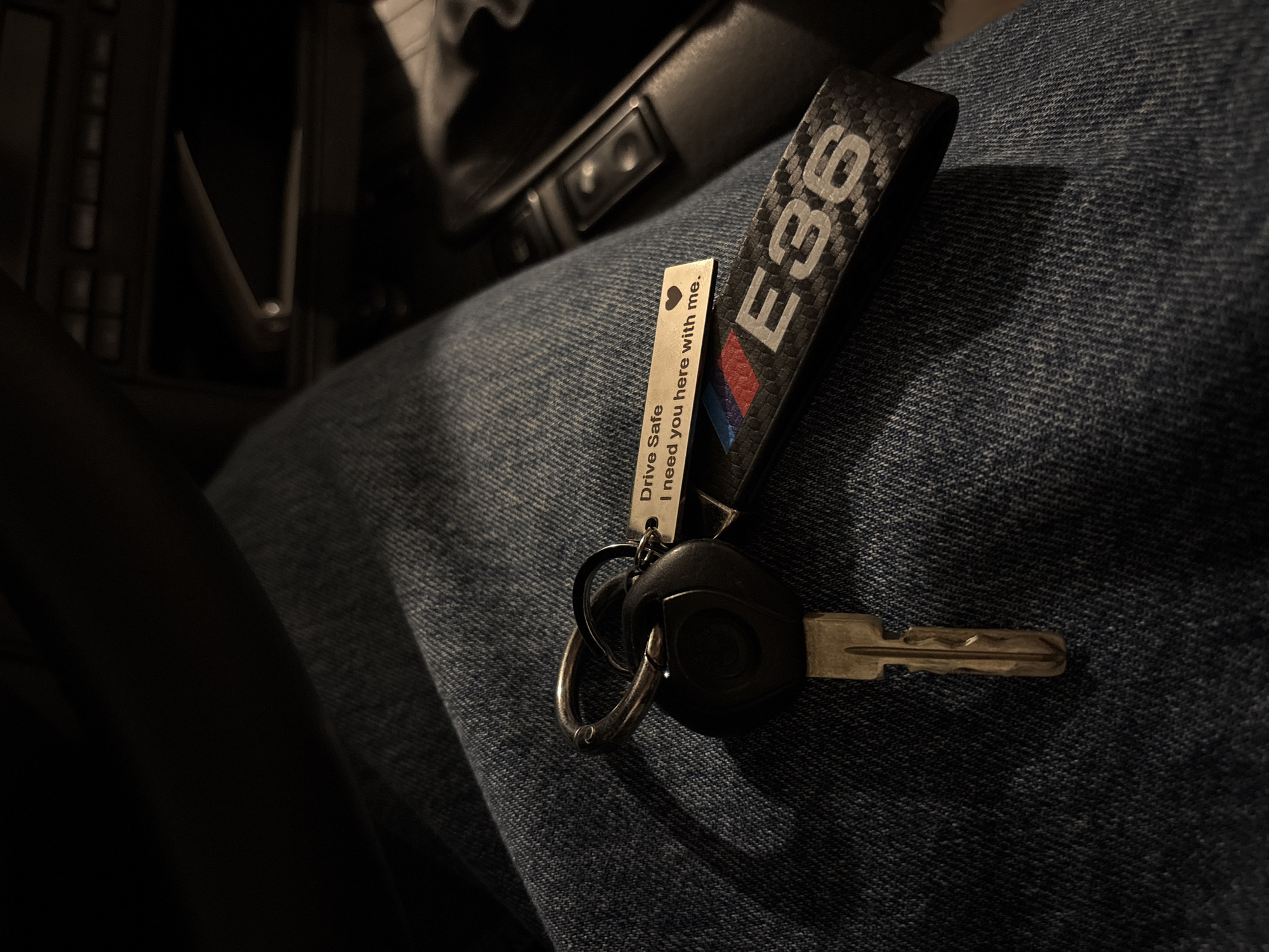 The image shows a set of car keys resting on a person's lap covered in denim. The keys have a black fob and a silver key. Attached to the keyring are two keychains: a black carbon fiber keychain with the text "E36" and the BMW M colors, and a silver keychain with the text "Drive Safe. I need you here with me." A gearshift and buttons are visible in the upper background.