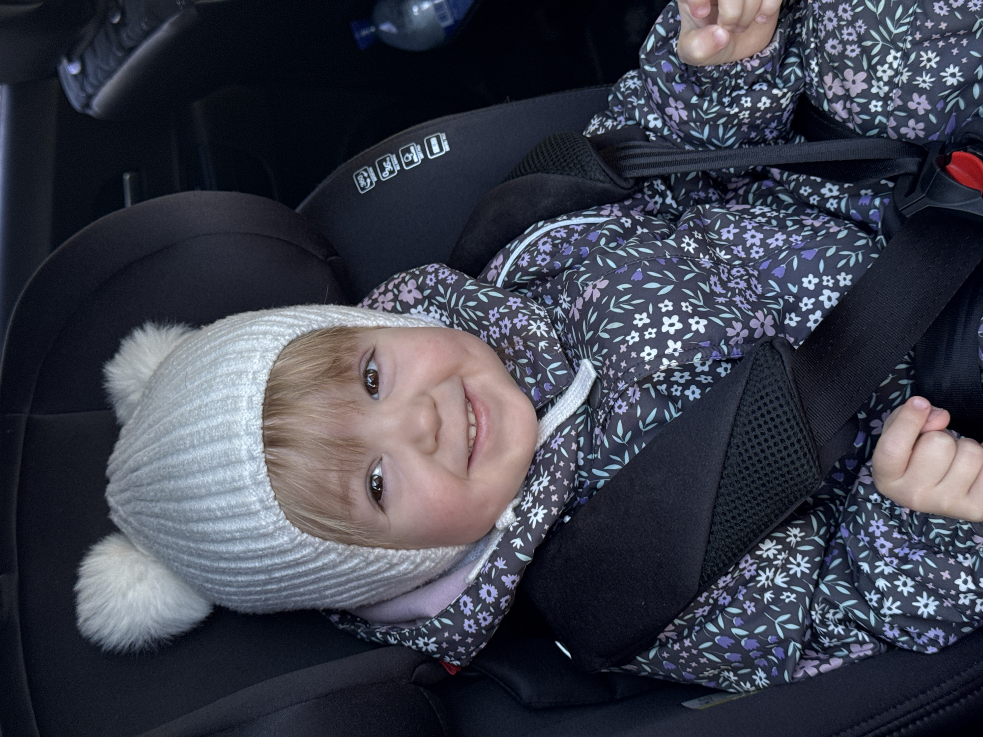 A young child wearing a white knit hat with two pom-poms and a floral patterned jacket is strapped into a black car seat. The child is smiling. In the background, a water bottle is visible. It is impossible to determine whether this photo was taken in Chaikovsky, Russia.