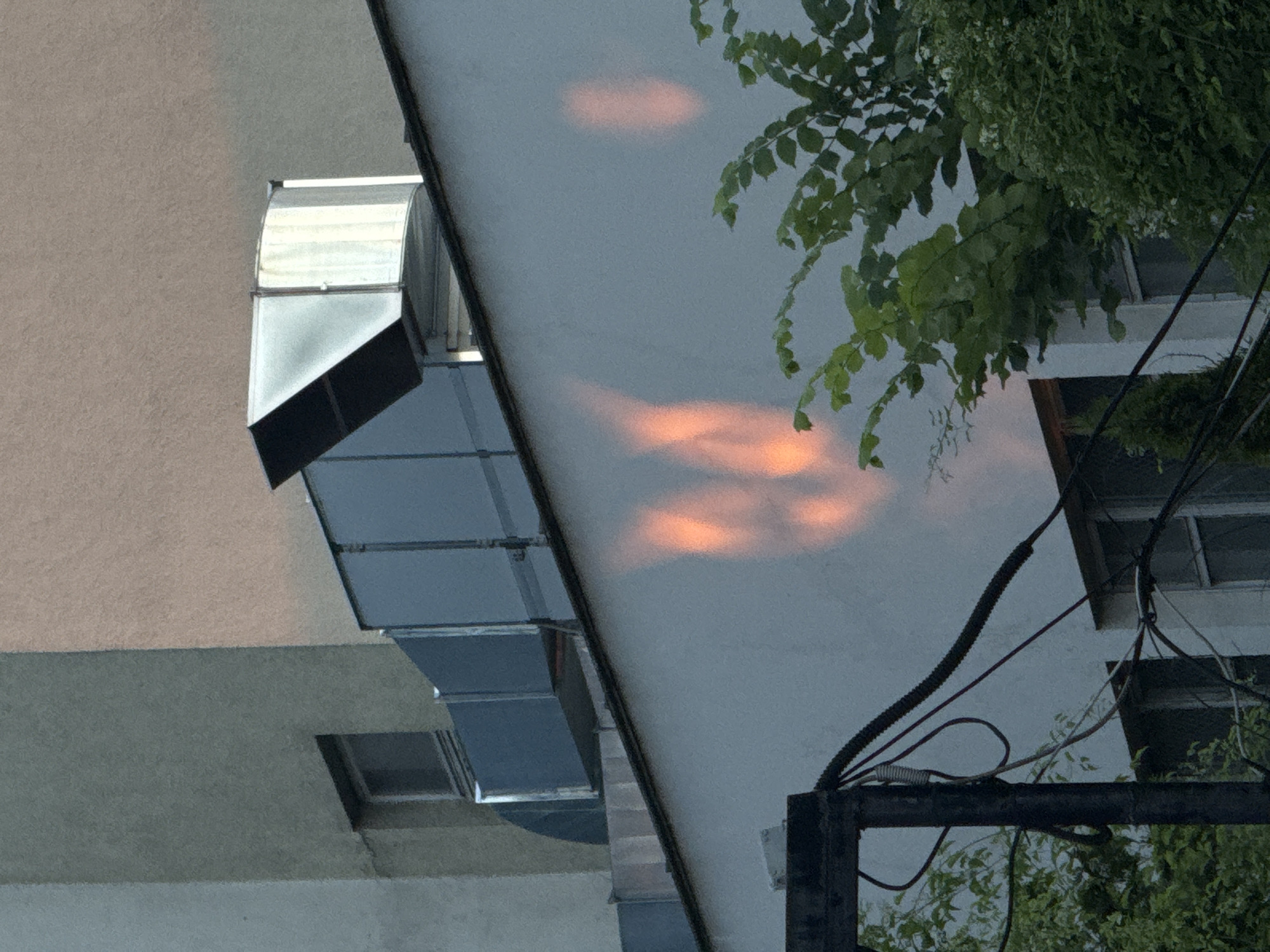 The image depicts an exterior view of a building's upper floors and rooftop in an urban environment, likely Sofia, Bulgaria. Prominent features include large, metallic ventilation ducts running along the rooftop, with a curved, angular section at the top right. The building's facade is primarily white, with visible windows below the rooftop level. A large section of the white wall in the foreground displays an abstract, orange-hued reflection or shadow, possibly cast by sunlight interacting with an unseen object or architectural element. To the right, lush green foliage, including trees and bushes, partially obscures the building. In the lower left quadrant, a dark metal structure with numerous electrical wires and conduits is visible. The sky above is overcast, with a gradient of lighter and darker gray tones.