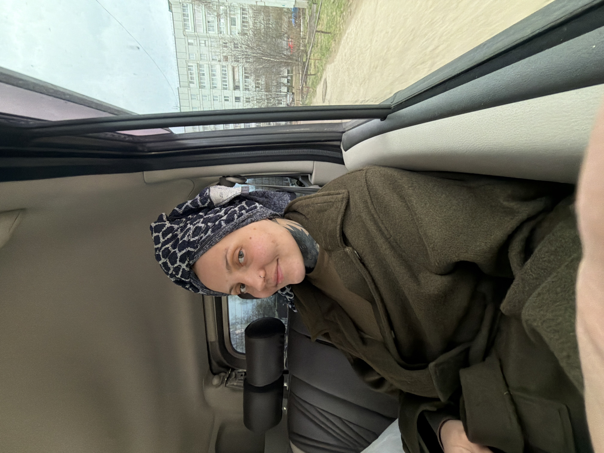 A young woman is depicted from the chest up, seated inside a vehicle and looking towards the camera with a soft, gentle smile. She wears a patterned black and white head covering, a dark olive green coat, and a dark long-sleeved garment underneath. A tattoo is partially visible on her left arm. She has light-colored eyes and a small nose piercing.
The setting is inside a car or similar vehicle, with a light-colored headliner and dark-colored seats visible. Through the vehicle's windows, an urban or suburban landscape in Chaikovsky, Russia, is visible. The background features several multi-story residential buildings, characteristic of post-Soviet architecture, under an overcast sky. Bare trees and sections of a dirt path or road with some grass are also present, suggesting the season is late autumn, winter, or early spring. The overall lighting is diffused and grey, indicating an overcast day. No text is visible in the image.