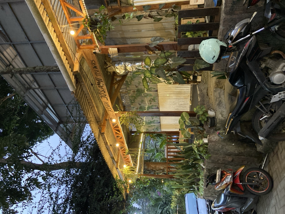 The image depicts the entrance area of "Kedai Josami Trawas," an establishment located in Cembor, Indonesia, at dusk or early evening. The building features a multi-tiered corrugated metal roof supported by wooden beams, with the name "KEDAI JOSAMI Trawas" painted in white lettering on the underside of the main canopy. String lights illuminate the ceiling structure. The entrance area is heavily adorned with various green potted plants and climbing foliage arranged on stone ledges and around wooden pillars, creating a natural aesthetic. Two motorcycles are parked in the foreground: a red and orange scooter (Honda Vario type) on the left, and a black sport motorcycle (Yamaha V-Ixion type) on the right, which has a light blue open-face helmet resting on its seat. A partial view of a light-colored car is visible behind the scooter. The ground surface in the immediate foreground is paved. Stone steps lead up from the parking area into the establishment. The background reveals dense trees and vegetation against an overcast or twilight sky. No individuals are present in the scene.