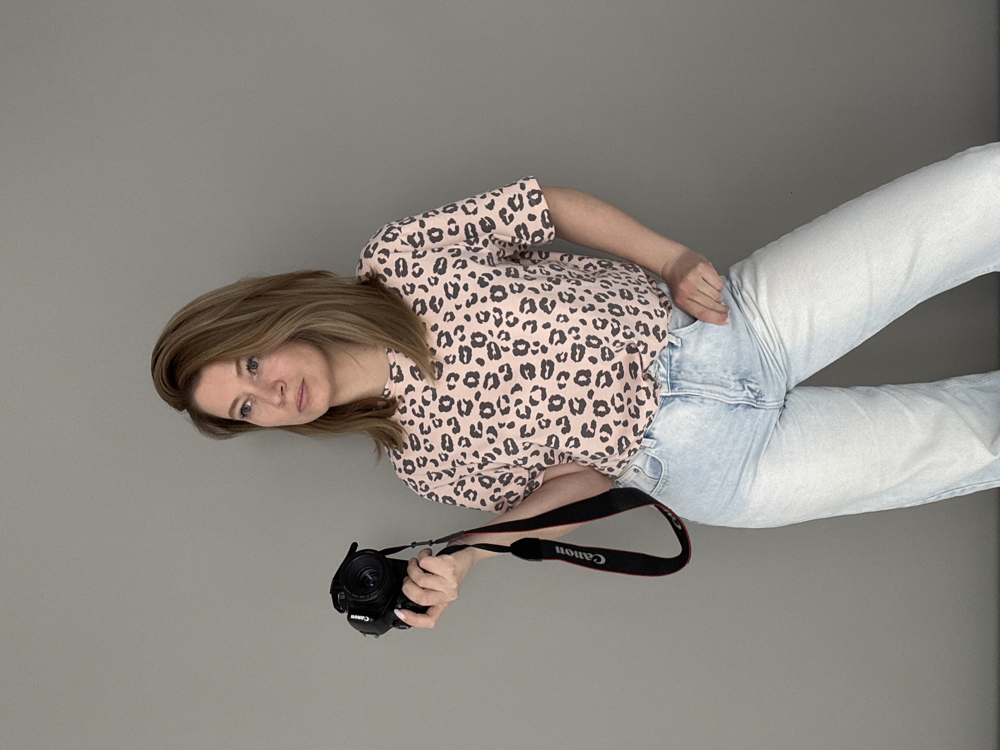 A woman with shoulder-length brown hair and light blue jeans stands against a gray background, holding a Canon camera with a black strap in her left hand. She wears a pink and black leopard-print shirt and looks toward the viewer. There is no indication of the city of Chaikovsky, Russia.