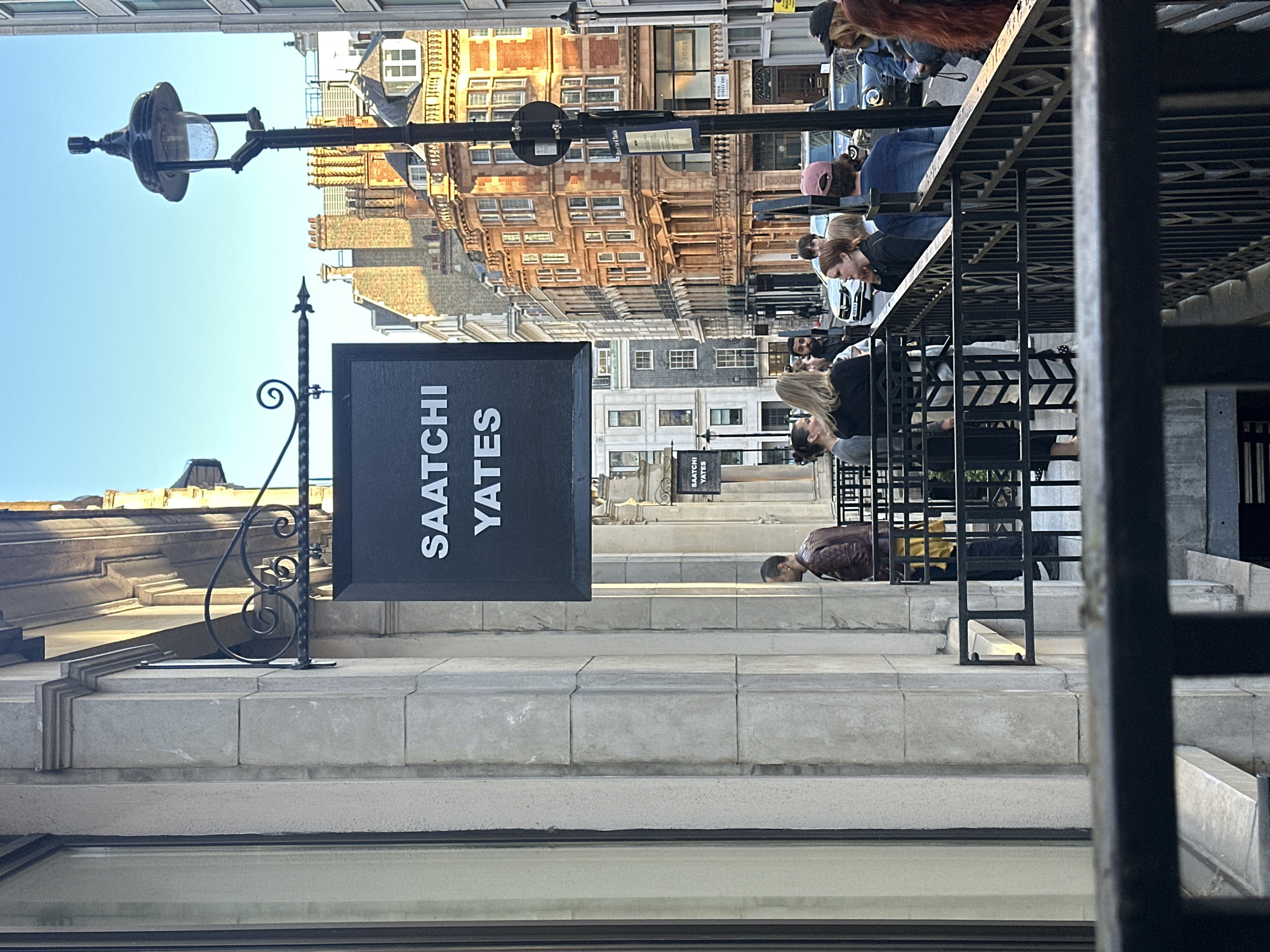A photograph, captured from a slightly elevated perspective, shows a scene from Saatchi Yates, a gallery located in London, United Kingdom. The gallery's dark-colored sign, emblazoned with the name "SAATCHI YATES" in white, is prominently displayed. A black lamppost is visible. The architecture of the buildings across the street is multi-story and features a variety of window designs and brick colors, indicating a mix of old and new construction. The sky is a clear light blue. Several individuals are present, including people sitting and standing on the steps of the gallery. Some people are looking at the steps or leaning forward.