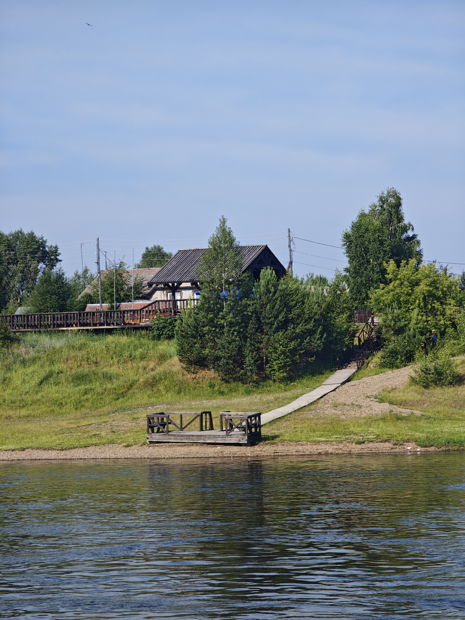 The image depicts a serene waterside scene in Galanino, Russia. In the foreground, a calm body of water reflects the sky and surrounding greenery. A small wooden dock juts out from the sandy bank into the water, equipped with simple seating. A paved path, possibly a walkway or ramp, leads from the bank upwards towards a slightly elevated area. The middle ground features a lush green grassy slope dotted with trees. A wooden boardwalk runs along the top of the slope, bordered by a railing. Behind the boardwalk, a cluster of houses with dark roofs and some exposed wooden structures is visible. Numerous trees, including birch and pine, are scattered throughout the landscape, some reaching considerable heights. The sky is a clear, pale blue with a few faint wispy clouds and a distant bird in flight. The overall impression is one of a peaceful, natural environment with some human infrastructure.