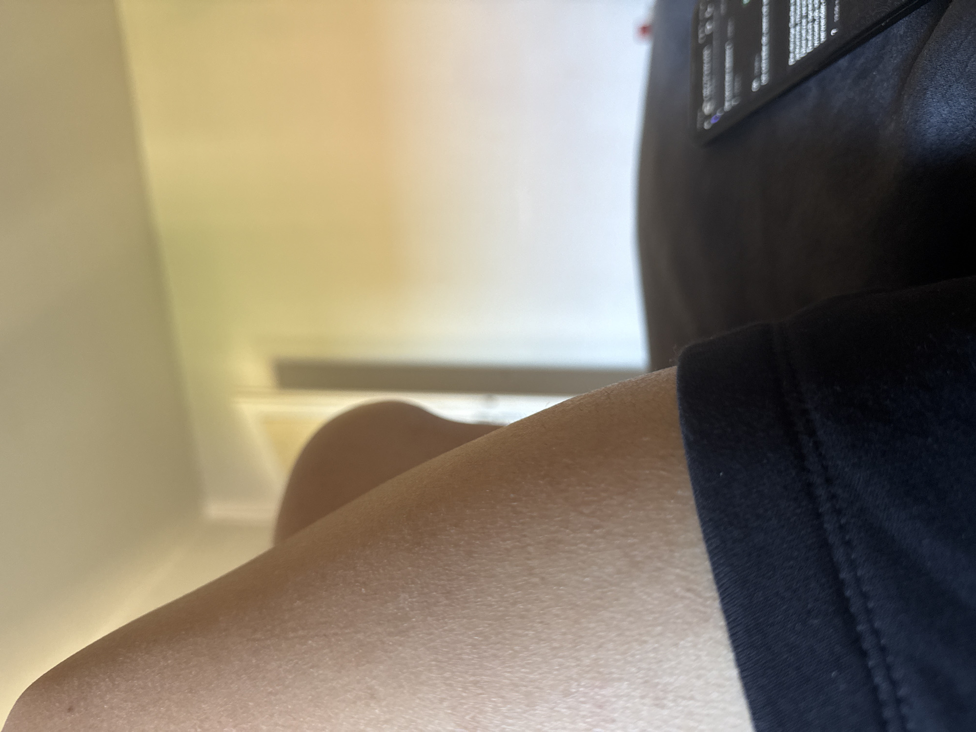 The image shows a close-up view of a person's leg and a black fabric, likely clothing, in the foreground. The leg appears to be light-skinned with visible pores and texture. In the background, a wall and a doorway are visible, suggesting an indoor setting. The lighting is warm and diffused, casting a soft glow on the wall. A dark object with illuminated text, possibly a smartphone or remote control, is partially visible on a dark surface in the lower right corner. The scene appears to be in Trimbach, Switzerland, based on the provided context, but there are no specific visual cues within the image to confirm this location.