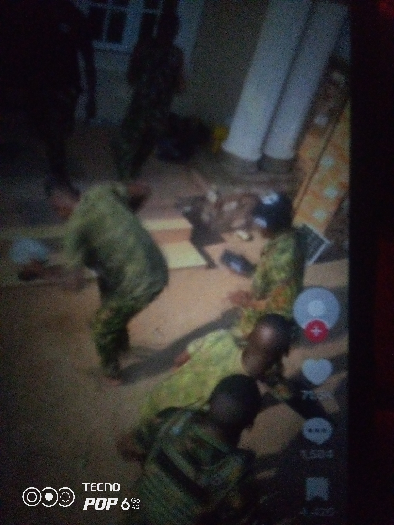 In an outdoor setting, likely a paved area with some uneven ground, three individuals are depicted. Two of the individuals are wearing camouflage-patterned clothing, suggesting a military or paramilitary context. One individual, positioned in the center, is wearing a green, yellow, and orange patterned shirt and a black helmet. Another individual to the left is wearing a similar camouflage outfit. The third individual, in the foreground on the right, is wearing a dark shirt and appears to be looking down. The scene appears to be captured during daylight, but the lighting is somewhat dim. A portion of a building with what looks like a white-framed window is visible in the background, as are two white cylindrical pillars. The overall image quality is low, with significant blur. A digital overlay is present on the right side of the image, featuring icons for likes, comments, and shares, along with numerical counts. There is also a watermark in the bottom left corner that reads "TECNO POP 6 Go 4G". The specific location in Howayi, Nigeria cannot be determined from the visual information provided.
