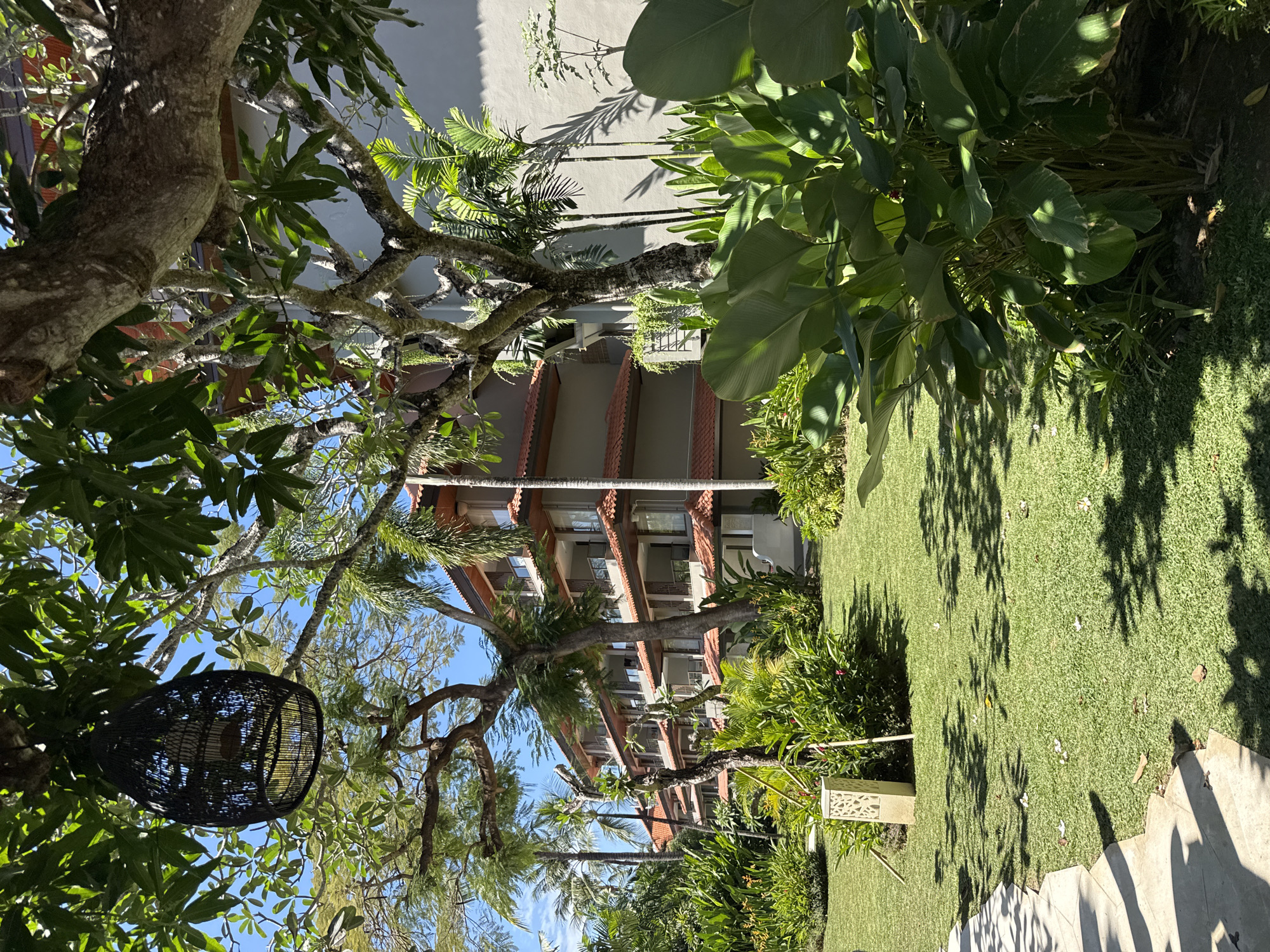 The image depicts a resort building and its surrounding tropical garden in Kuta Selatan, Indonesia. The building is a multi-story structure with a gray facade and red-tiled roofs, featuring balconies and numerous windows. Lush green vegetation, including large leafy plants, palm trees, and flowering bushes, dominates the foreground and sides of the image. A stone pathway is visible in the lower left, leading into the garden. In the upper left, a black woven lantern hangs from a tree branch. The ground is covered in manicured green grass, with shadows cast by the trees. The sky is clear and blue. There are no people visible in the scene.