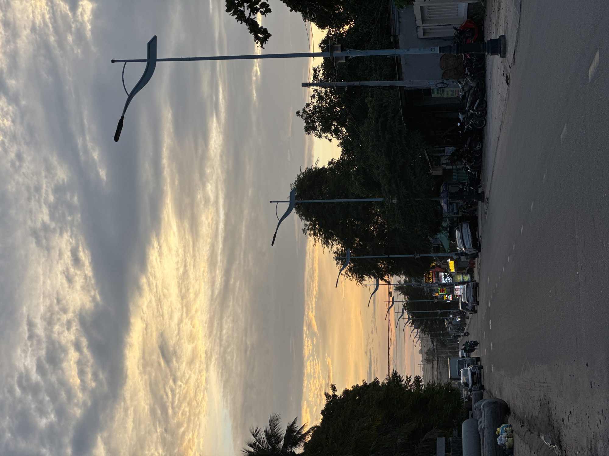 This vertical image captures a street scene at dusk in Mui Ne, Vietnam. The sky is filled with dramatic clouds, tinged with orange and yellow from the setting sun, creating a striking contrast with the darker gray clouds. A modern lamppost with a distinctive curved arm stands prominently in the foreground, silhouetted against the sky.
The street itself appears to be a main thoroughfare, with several cars and a motorcycle visible. On the left side of the road, a palm tree is visible, along with large concrete pipes and piles of trash near the curb. The right side of the street features a row of shops and buildings, with several motorcycles parked along the sidewalk. Trees and foliage line the street on both sides, and a smaller lamppost is visible further down the road. The overall atmosphere is that of a coastal town winding down for the evening.