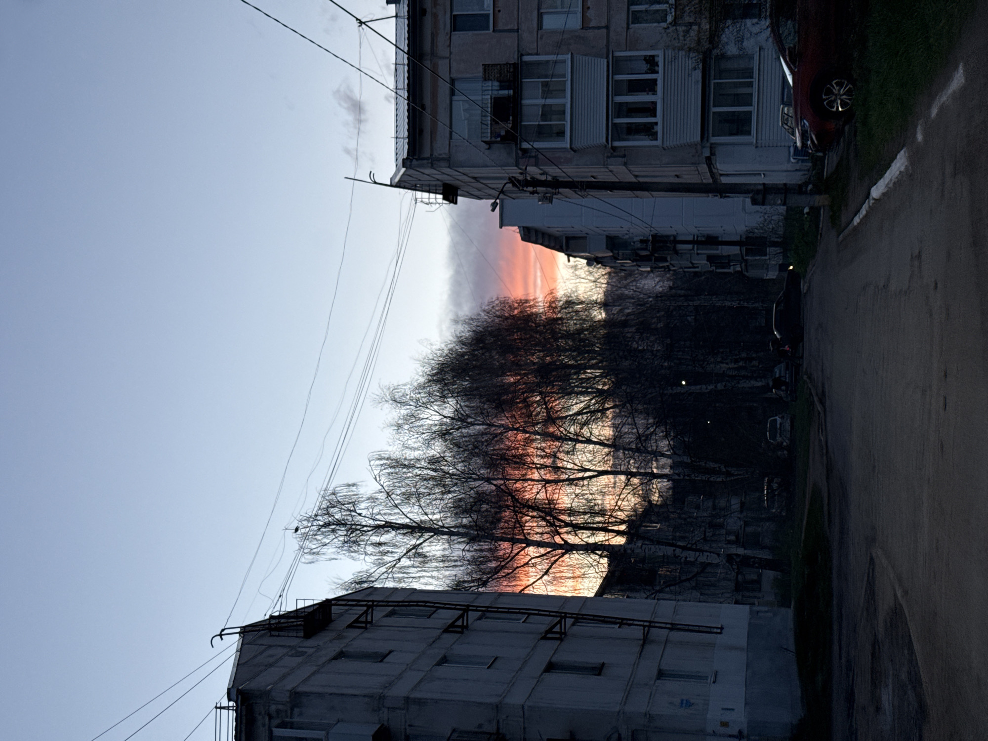 The image captures a street scene in an urban environment. Buildings frame the view of the sky, where a dramatic sunset with red and orange hues is visible through the branches of trees. Power lines stretch across the sky between the buildings. The street itself is dimly lit. A dark-colored car is parked on the right side of the street, adjacent to a grassy area. The buildings are multi-story and appear to be older residential structures.