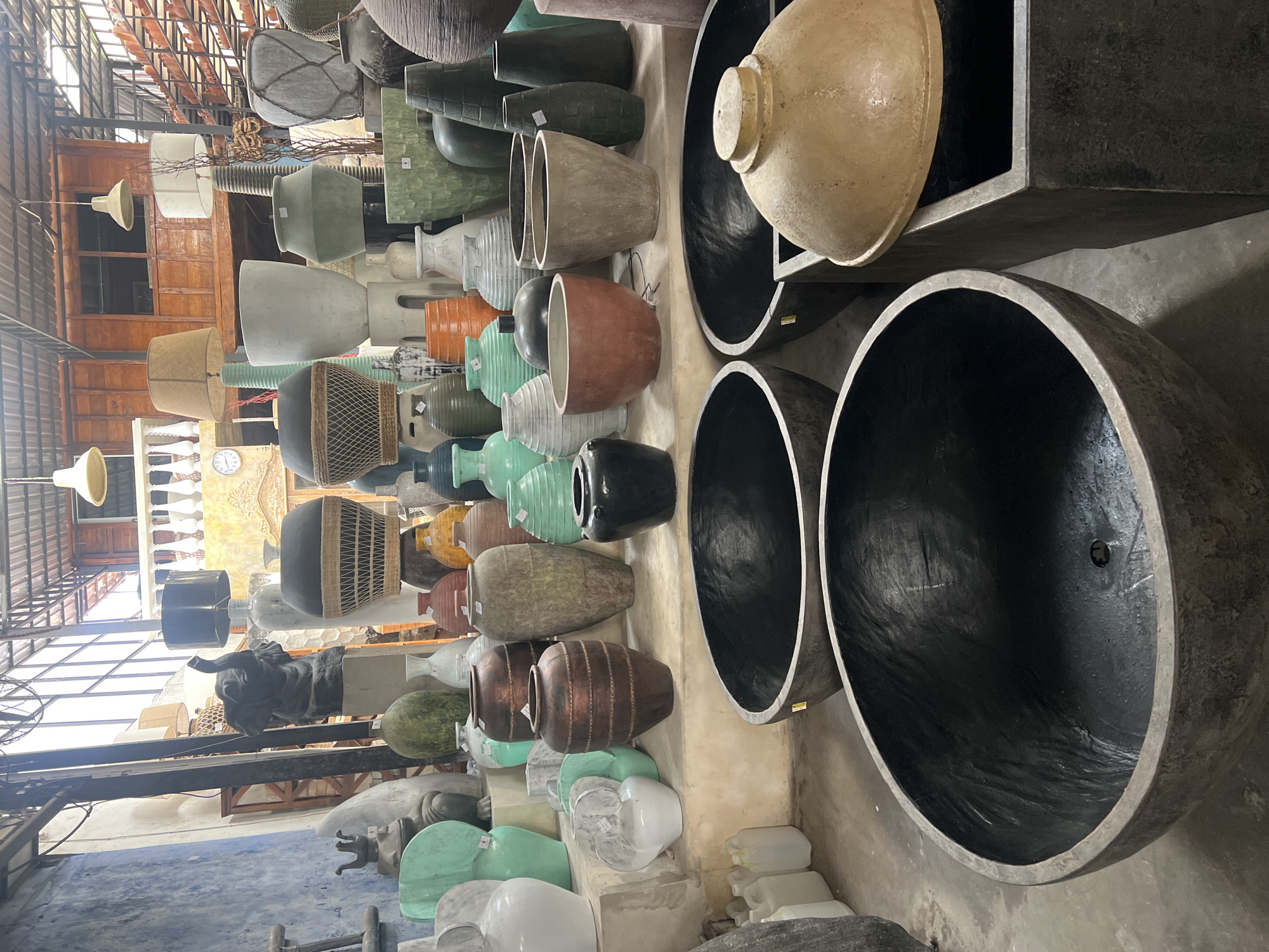 The image depicts a retail space, likely a pottery or garden decor shop, located in North Kuta, Indonesia. The environment is characterized by an open-air structure with a corrugated metal roof, allowing natural light to illuminate the scene. The foreground is dominated by several large, dark, basin-like concrete planters. Behind these, a variety of ceramic and concrete pots, vases, and decorative items are arranged on shelves and on the ground. These items vary in size, shape, and color, with prominent examples including large grey, green, and white vases, as well as smaller, intricately patterned pots. A notable feature is a large, dark grey, elephant-shaped sculpture positioned on a pedestal. The background reveals more of the shop's interior, including wooden structures, hanging lamps, and more shelving filled with decorative objects. No people are visible in the image.