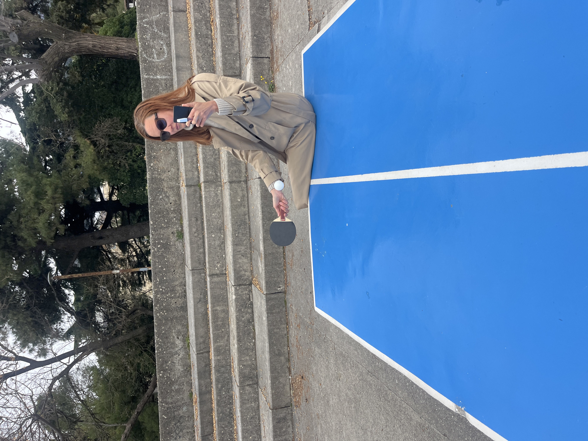 A woman in a tan trench coat and sunglasses stands on the edge of a blue outdoor ping-pong table next to concrete steps in Budva, Montenegro. She is holding a table tennis paddle and a white ping-pong ball, seemingly preparing to play. In her other hand, she holds a dark-colored smartphone. The background shows trees and vegetation typical of the Montenegrin coast. The scene is well-lit, suggesting daylight conditions.