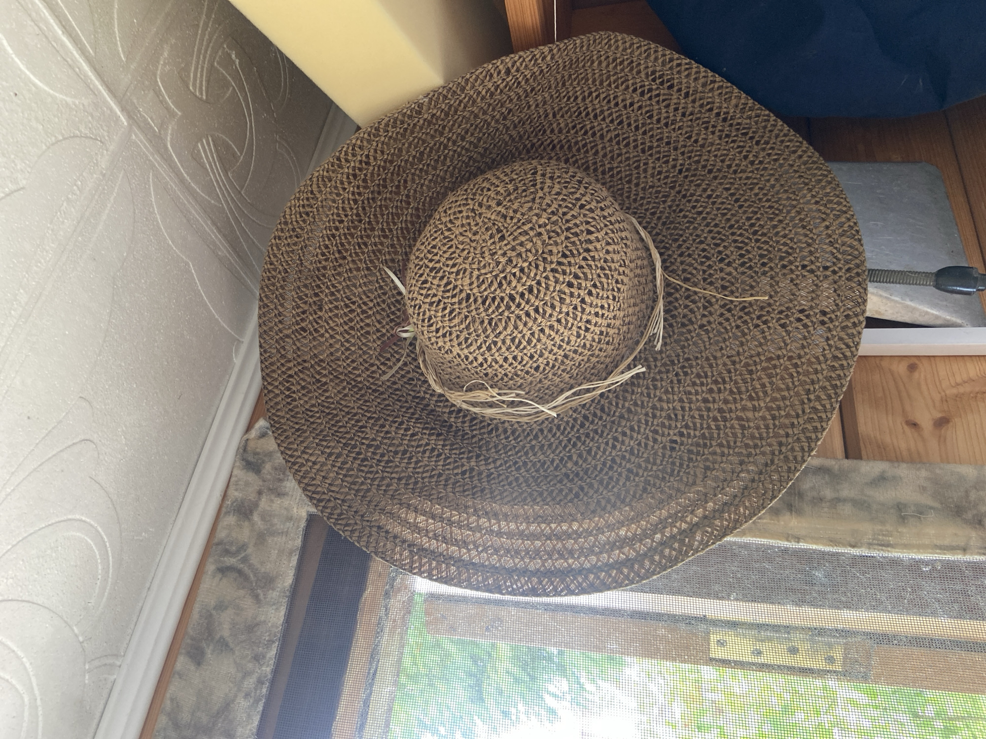 The image presents a close-up, overhead view of a large, wide-brimmed straw hat, likely a sun hat, made of light brown woven material. Several thin, lighter-colored straw or raffia strings are loosely tied around the base of the hat's crown.
The hat is positioned indoors, with various elements of the surrounding environment visible. To the left, a white wall features a distinct raised or embossed pattern of intertwined, curvilinear designs, resembling Celtic knots or a stylized floral motif. At the top of the frame, a segment of a light yellow or beige object is visible. On the right, wooden planks or panels are present, along with a dark blue object partially obscured in the upper right corner. Below this, a grey rectangular object with a dark, coiled spring or wire extending from it is visible, resting on a surface.
The bottom portion of the image shows a window with a dark mesh screen. Through the screen, bright daylight illuminates green foliage, suggesting trees or bushes outside. A wooden window frame and a metallic hinge are also noticeable. The scene depicts a static arrangement of objects in an indoor setting with a view to a sunny outdoor environment. There are no people, activities, or visible text in the image.