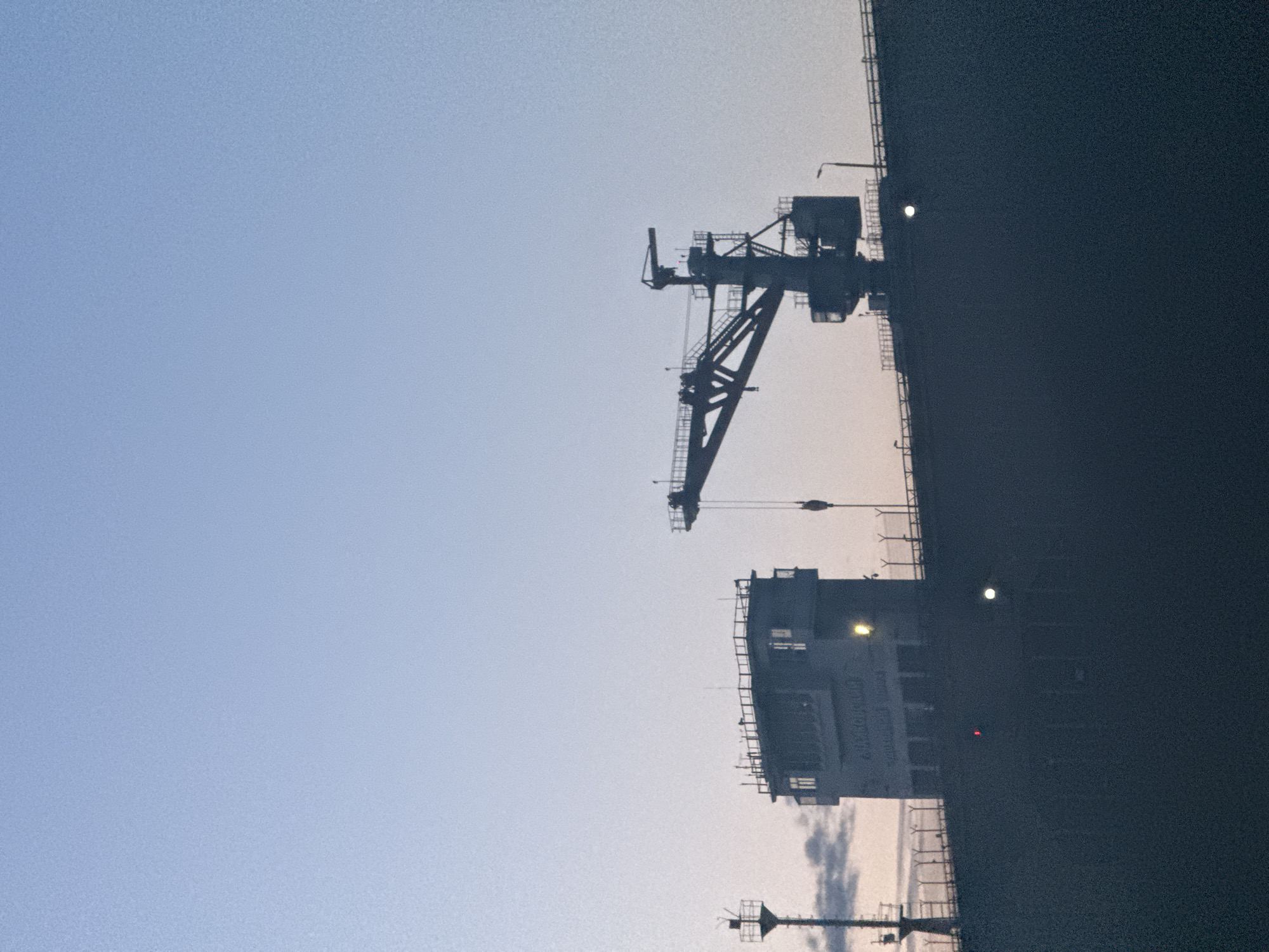 The image captures a large vessel, likely a cargo ship or industrial barge, docked or anchored in a port in Chaikovsky, Russia. The scene is set at either dawn or dusk, with the sky displaying a soft gradient from a light blue at the top to a paler, warmer tone near the horizon.
A prominent industrial crane, characterized by its complex metal framework, is silhouetted against the sky, suggesting loading or unloading operations. Below the crane, the ship's superstructure, containing the bridge or command center, is also silhouetted. Several artificial lights are visible on the ship's deck and superstructure, illuminating parts of the vessel in the dim light. The right side of the image is dominated by the dark mass of the ship's hull or cargo area.
On the side of the ship's superstructure, faintly visible text in Cyrillic characters reads "ЧАЙКОВСКИЙ" (Chaikovsky), confirming the vessel's name or its connection to the city. Below this, further indistinct text likely specifies "PORT" or "RIVER" and "RUSSIA". The sky is mostly clear, with a few faint wisps of clouds near the horizon. The overall impression is one of an active but quiet industrial maritime setting.