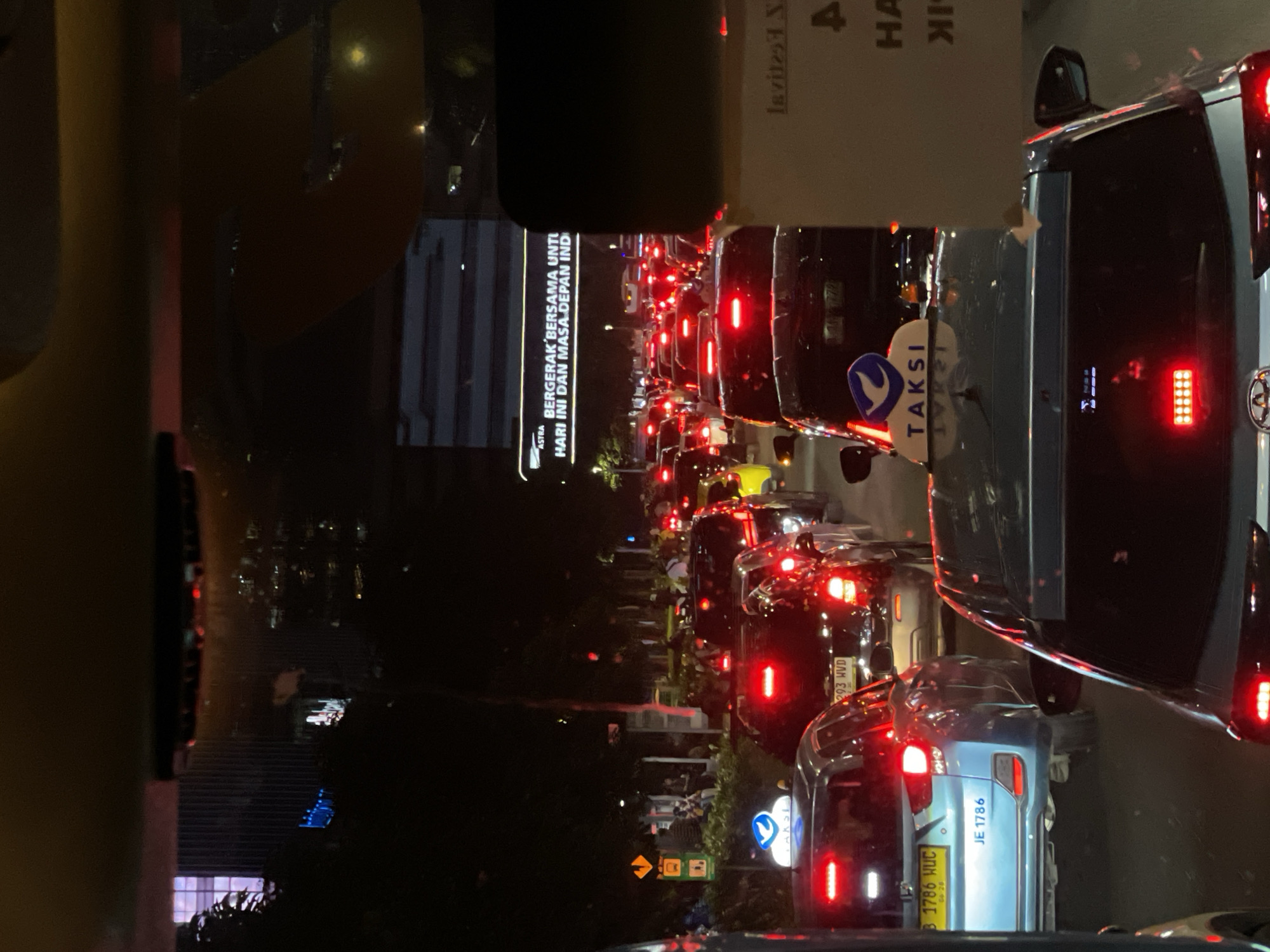The image shows a traffic jam at night in Central Jakarta, Indonesia. A long line of cars with illuminated taillights extends into the distance. There is a large advertising sign that reads "ASTRA BERGERAK BERSAMA UNTU HARI INI DAN MASA DEPAN IND". A taxi is also seen with a "TAKSI" sign. The car in the foreground has license plate "JE 1786" and another one has the license plate "B 1786 HUC". Buildings are visible in the background.