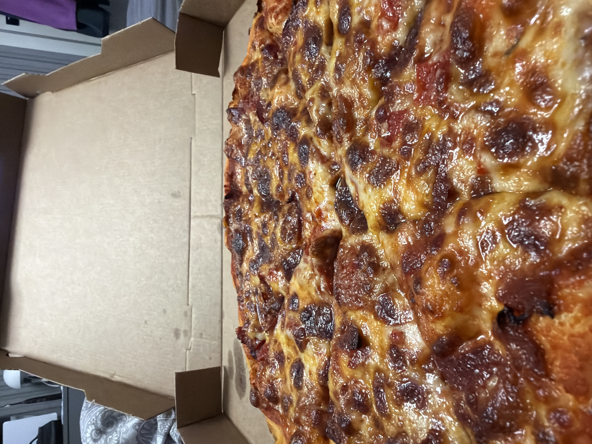 The image shows a close-up of a pizza inside an open cardboard pizza box. The pizza is topped with melted cheese and what appears to be pepperoni or a similar meat. The crust is golden brown and appears slightly crispy. The pizza box is light brown cardboard and is open, with the lid propped up. The background is slightly out of focus, but shows a bed with a patterned comforter and what looks like a wall or furniture. There are no people visible in the image. The scene is indoors. There is no information about the location in Ruse, Bulgaria.