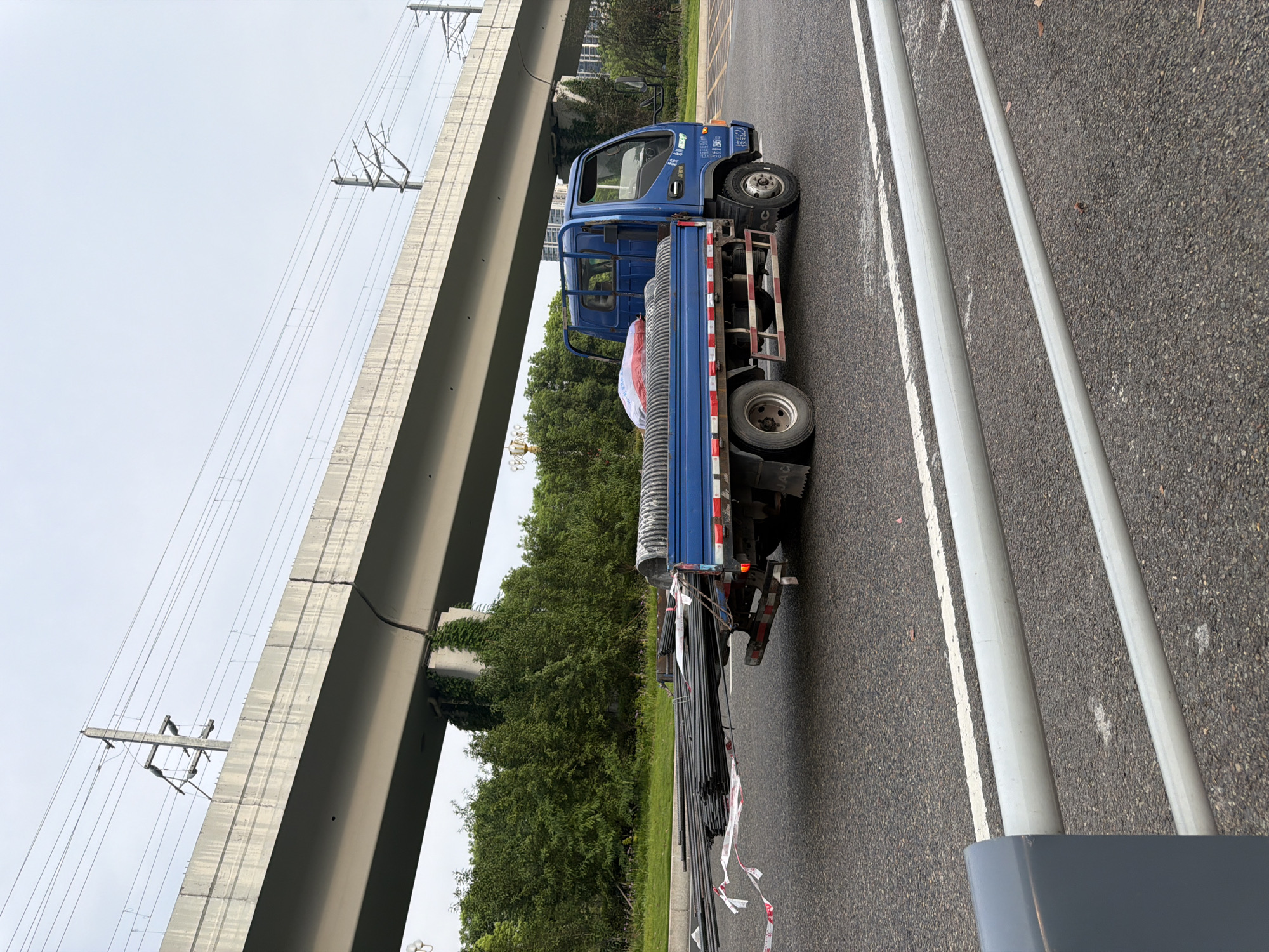 A blue flatbed truck is parked on a paved road, under a raised concrete structure that appears to be part of an elevated highway or train line, in the city of Jiangjin, China. The truck is loaded with long, dark metal rods, some of which extend past the back of the truck and are secured with red and white caution tape. There are also some cylindrical objects wrapped in plastic on the truck bed. Trees and vegetation are visible behind the truck. The sky is overcast. The scene is captured from a low angle, slightly behind and to the right of the truck, focusing on the vehicle and its cargo.