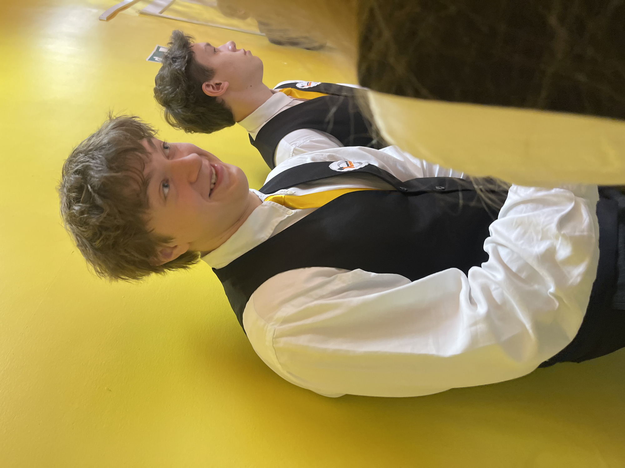 The image shows two young men in formal attire, consisting of white shirts, black vests, and yellow ties, lying on a yellow floor. The location might be in Krasnoyarsk, Russia, as suggested in the prompt, but no specific elements in the photo confirm this. The foreground figure smiles directly at the camera. The background figure is further away and appears to be looking upwards. Both are wearing badges.