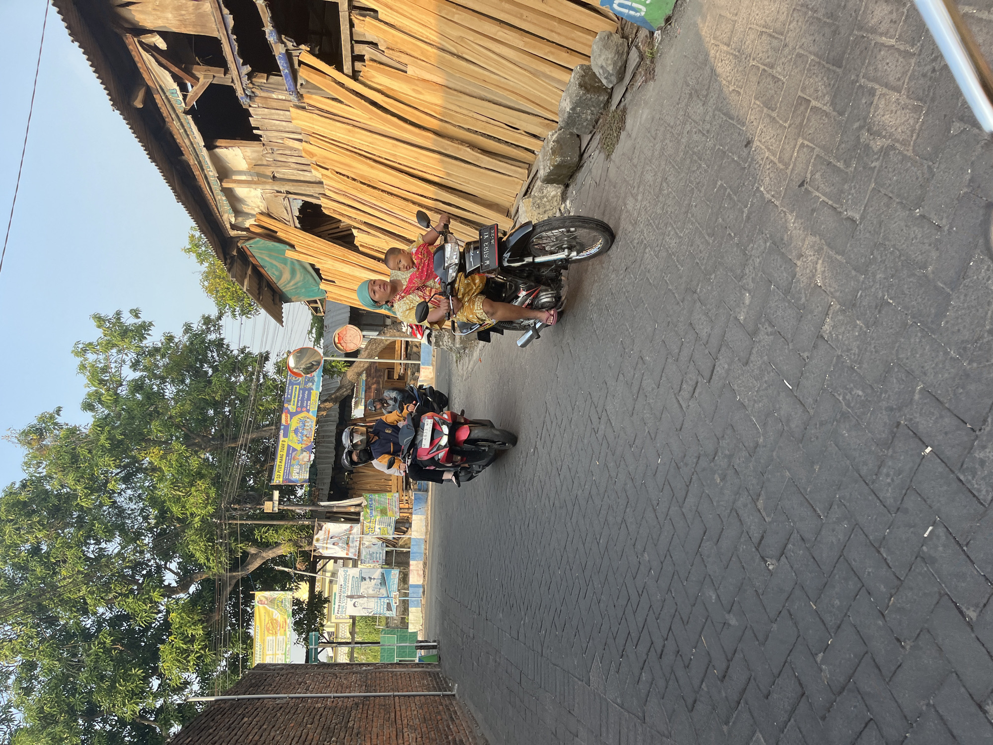 The image depicts a street scene in Tenggulunan, Indonesia, likely in a semi-urban or rural area, characterized by a brick building on the left and a structure made of stacked wooden planks on the right. Two motorcycles are the primary focus of activity. In the foreground, a woman wearing a blue headscarf and a patterned yellow and red outfit rides a black motorcycle, carrying a baby on her lap. The baby is wrapped in a red cloth. The woman wears red flip-flops. In the mid-ground, another motorcycle, red in color, is ridden by two individuals wearing helmets. The rider is wearing a jacket with yellow and blue accents, and the passenger appears to be wearing a dark top. A third motorcycle, partially visible in the background on the right, is ridden by a person wearing a helmet. The street is paved with dark interlocking bricks, laid in a herringbone pattern. The environment includes trees with green foliage and several banners or signs displayed on poles along the street, some with text and images, suggesting a commercial or public area. The lighting indicates it is daytime, possibly late afternoon, given the long shadows. The overall atmosphere is one of daily life and transit in a local Indonesian setting.