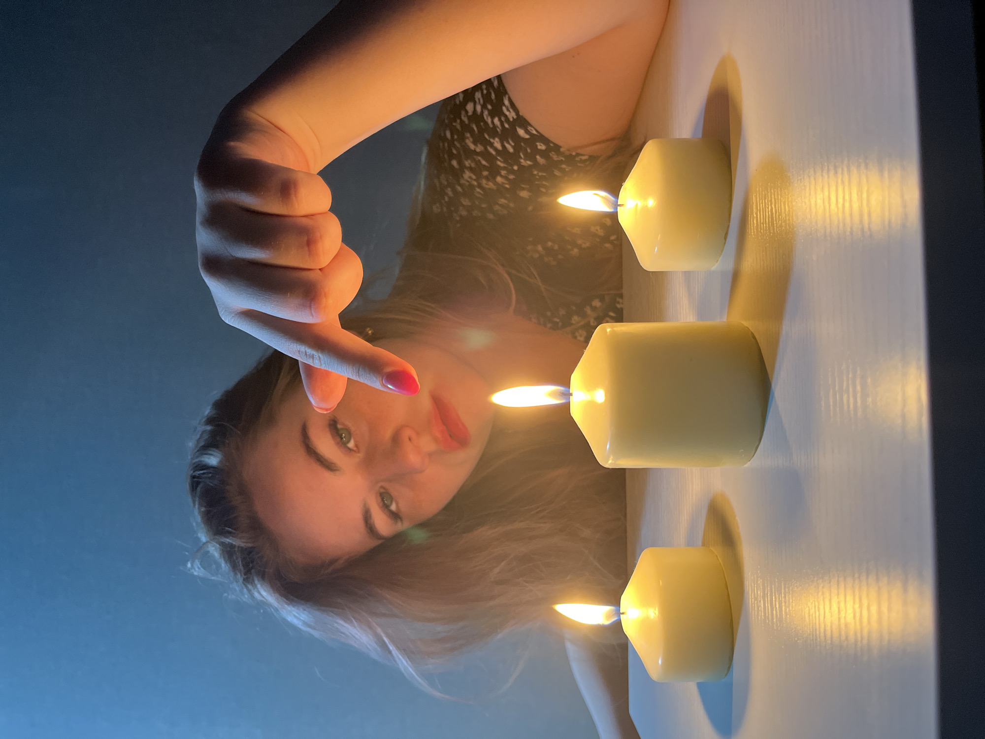 An intimate, low-angle, close-up image shows a young woman looking directly at the viewer, positioned beneath three lit candles. Her face, with green eyes and lips adorned with red lipstick, is partly obscured by her long, light brown hair. She is wearing a dark top with a subtle light pattern. Her right hand, featuring red nail polish on her index finger, is extended and pointing towards the candles.
Three cream-colored, pillar-style candles are arranged vertically on a light-colored, flat surface to the right of the frame. Each candle is lit, casting warm, elongated shadows on the surface. The flames provide the primary illumination for the scene, creating a soft, warm glow. The background behind the woman is a muted, dark blue, suggesting an indoor setting in low light conditions. The overall atmosphere is contemplative and warm, likely taking place in the evening or at night. No text is visible. While the location context is Chaikovsky, Russia, there are no specific visual cues in the image itself to confirm this.