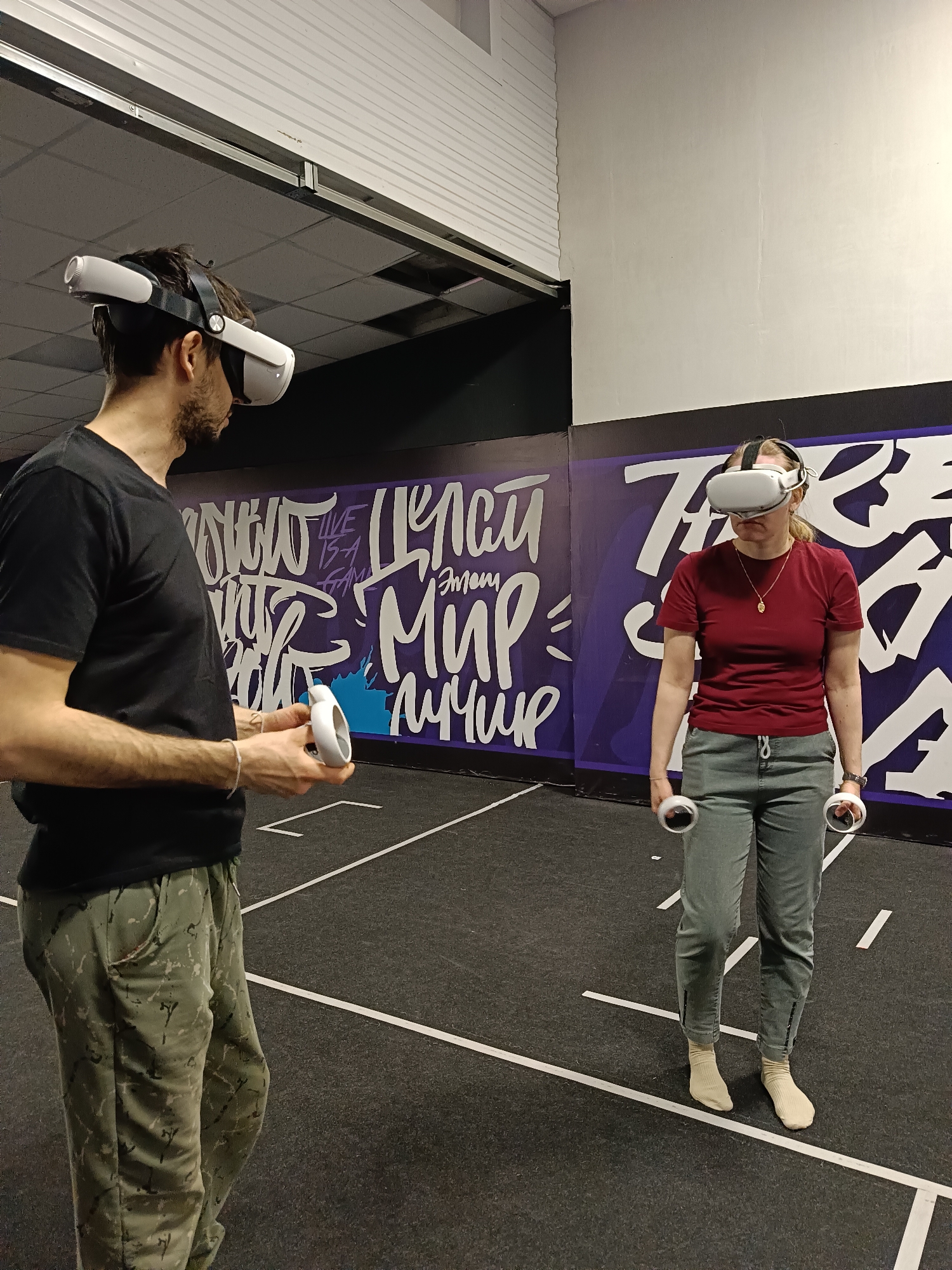 Two individuals, a man and a woman, are engaged in a virtual reality experience within an indoor setting. Both are wearing white VR headsets, likely Meta Quest 2, and holding matching white controllers. They stand on a dark gray floor marked with white lines, suggesting a dedicated play area.
The man, positioned on the left, is seen from the side, wearing a black t-shirt and camouflage-patterned pants. He holds one controller in his right hand, looking towards the right side of the frame. The woman, standing to the right and facing forward, wears a maroon t-shirt, gray-green pants, and light-colored socks without shoes. She holds a controller in each hand, her gaze directed straight ahead, obscured by the headset.
The background features a vibrant purple wall adorned with white, graffiti-style text. Visible phrases include "LIVE IS A GAME" and Russian words that translate to "MAKE THIS WORLD BETTER" (СДЕЛАЙ ЭТОТ МИР ЛУЧШЕ). The upper section of the wall transitions to a lighter color. The ceiling is industrial, with a corrugated metal section and visible structural elements.
The scene depicts a modern entertainment or gaming venue, likely a VR arcade or club, in Chaikovsky, Russia. The focused postures of the participants suggest immersive engagement in the virtual reality environment.