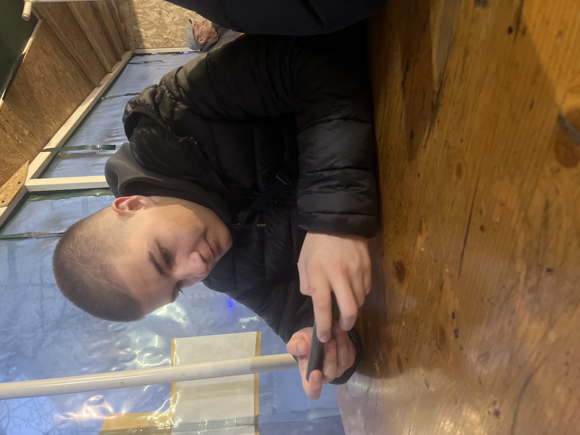 A young person with a shaved head, wearing a black puffer jacket, is holding a small, dark object in their hands. They are leaning over a wooden surface. Behind them, a structure with white rails and a semi-transparent, light blue material is visible. In the background, there is wooden construction.
The location is Asenovgrad, Bulgaria.